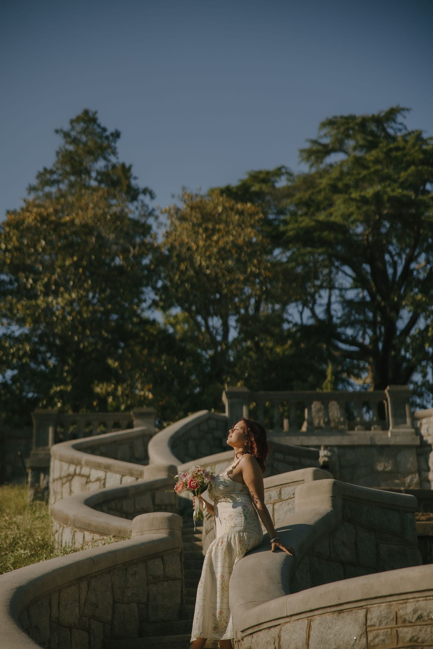 Woman in white dress holding bouquet stands on curved stone steps surrounded by lush green trees in golden sunlight.