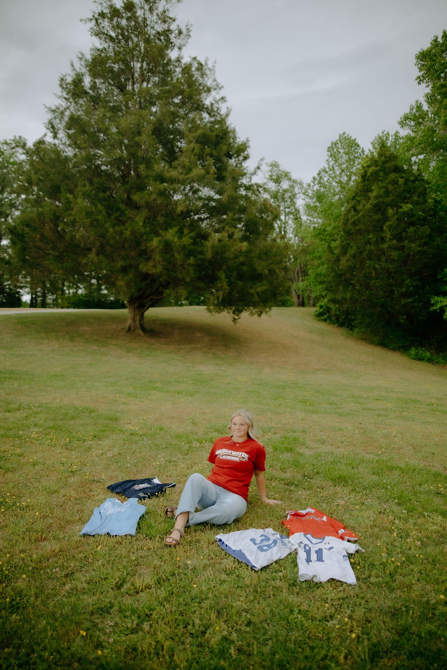 Woman in red shirt sits on grass surrounded by sports jerseys in a scenic park with lush green trees.