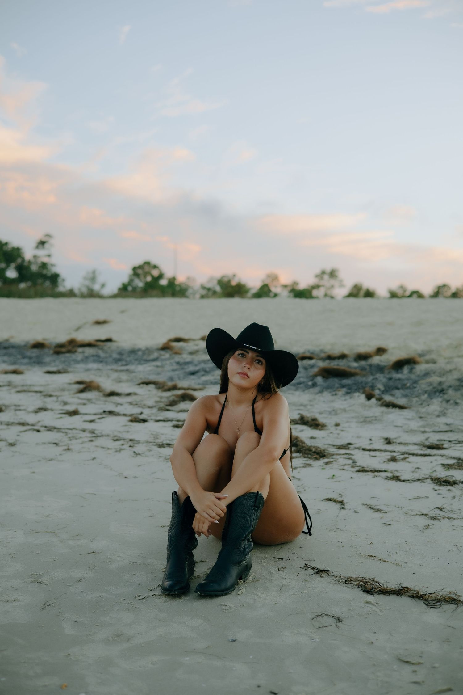 Woman in black cowboy hat and boots sitting on sandy beach at sunset.