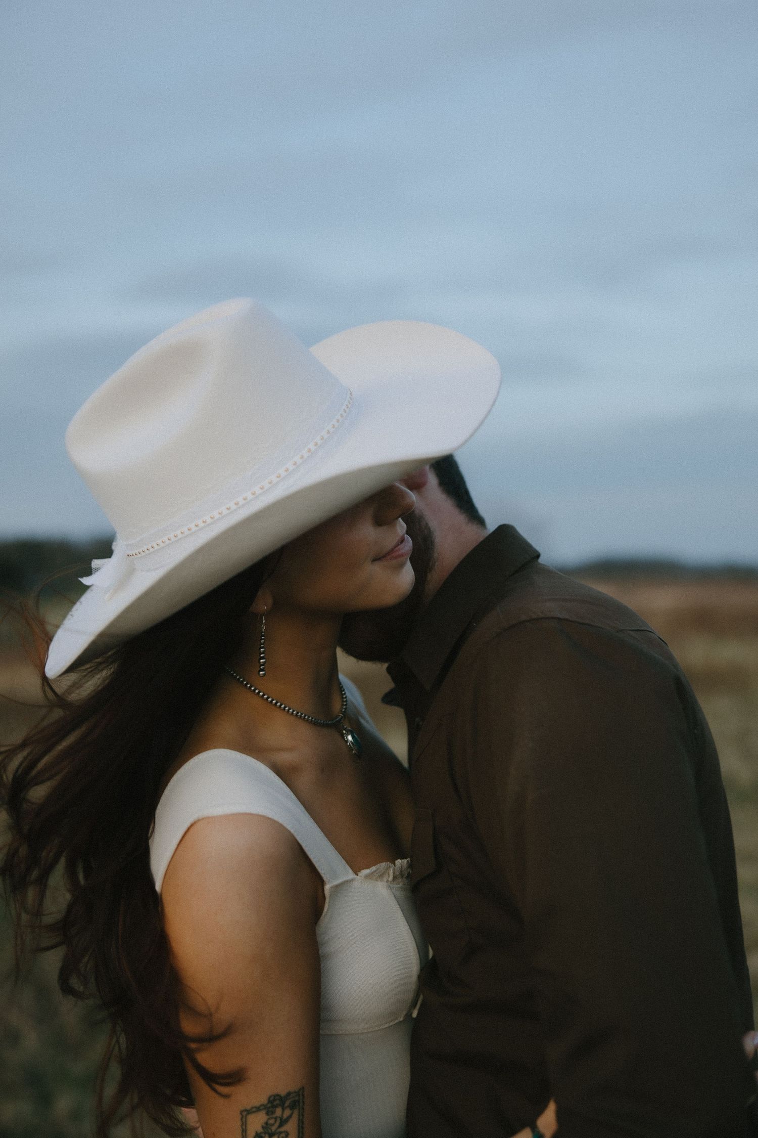 A bride in a white cowboy hat and dress shares a romantic moment with her groom in a rustic outdoor setting.