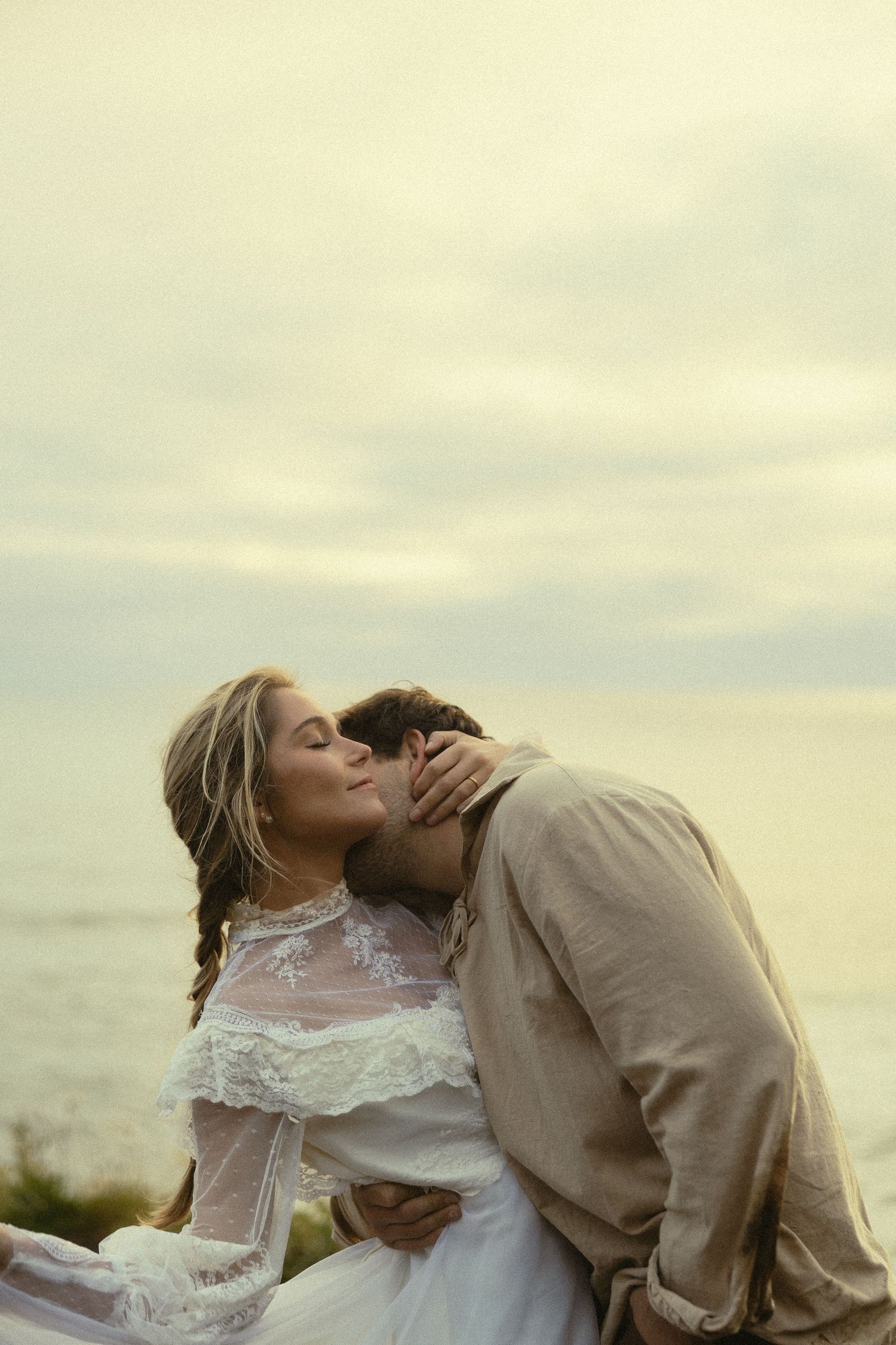Groom kisses bride in lace wedding dress by the ocean at sunset in a tender romantic embrace.