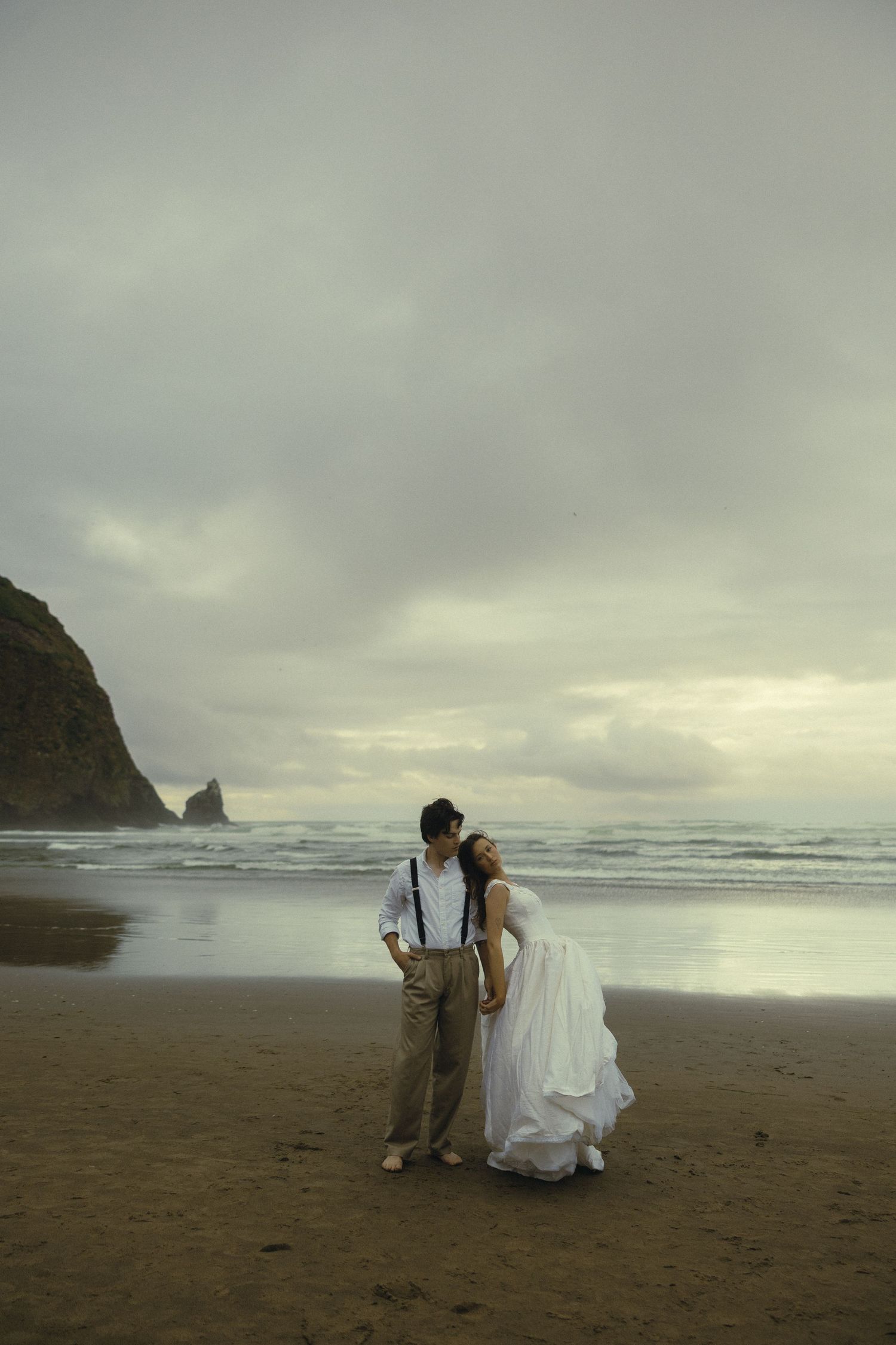 Couple in wedding attire embracing on a moody beach with rocky cliffs and dramatic cloudy skies.
