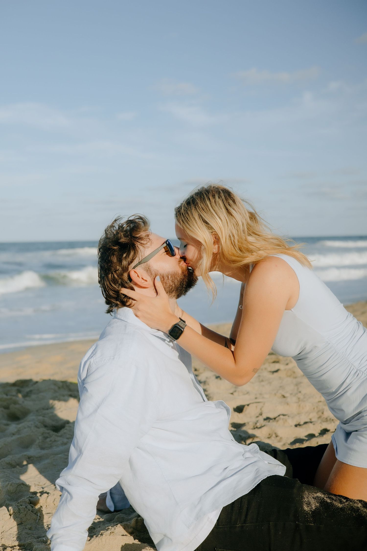 Two people share a romantic moment on a sandy beach with ocean waves in the background.