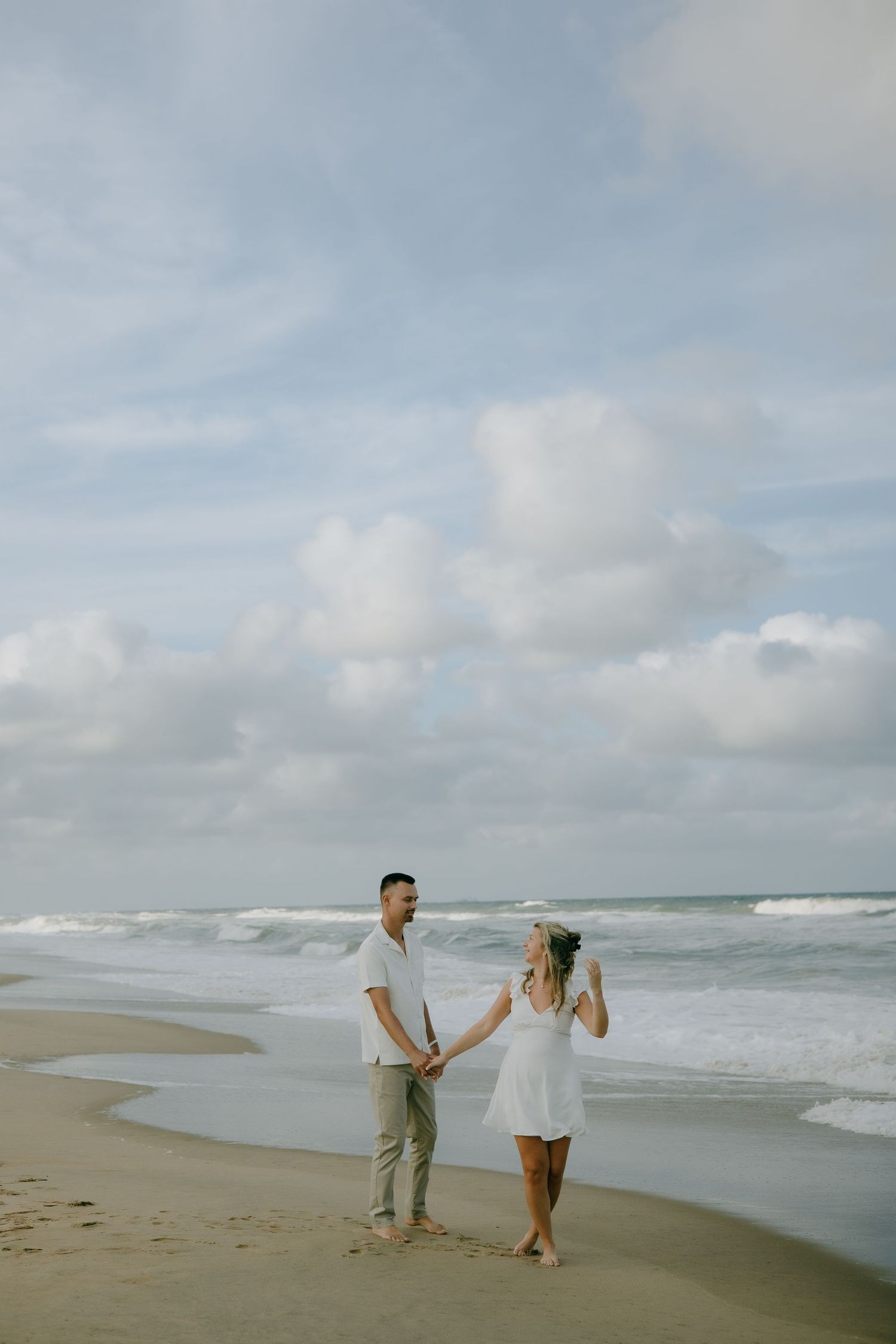 Two people playfully dance together on a serene beach with waves in the background.