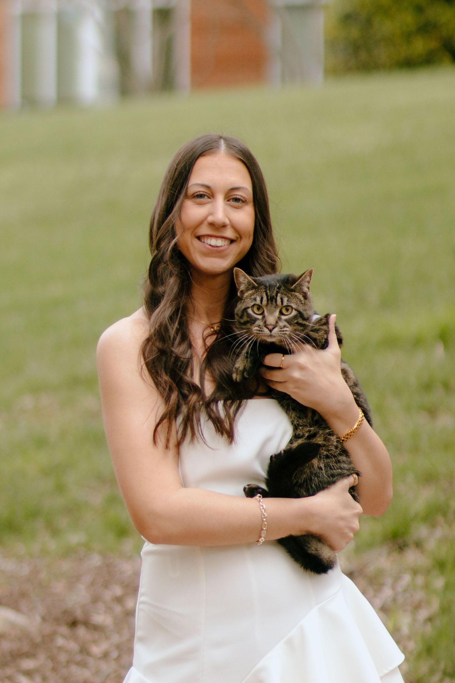 A person in white sits on grass while holding and smiling at a tabby cat.
