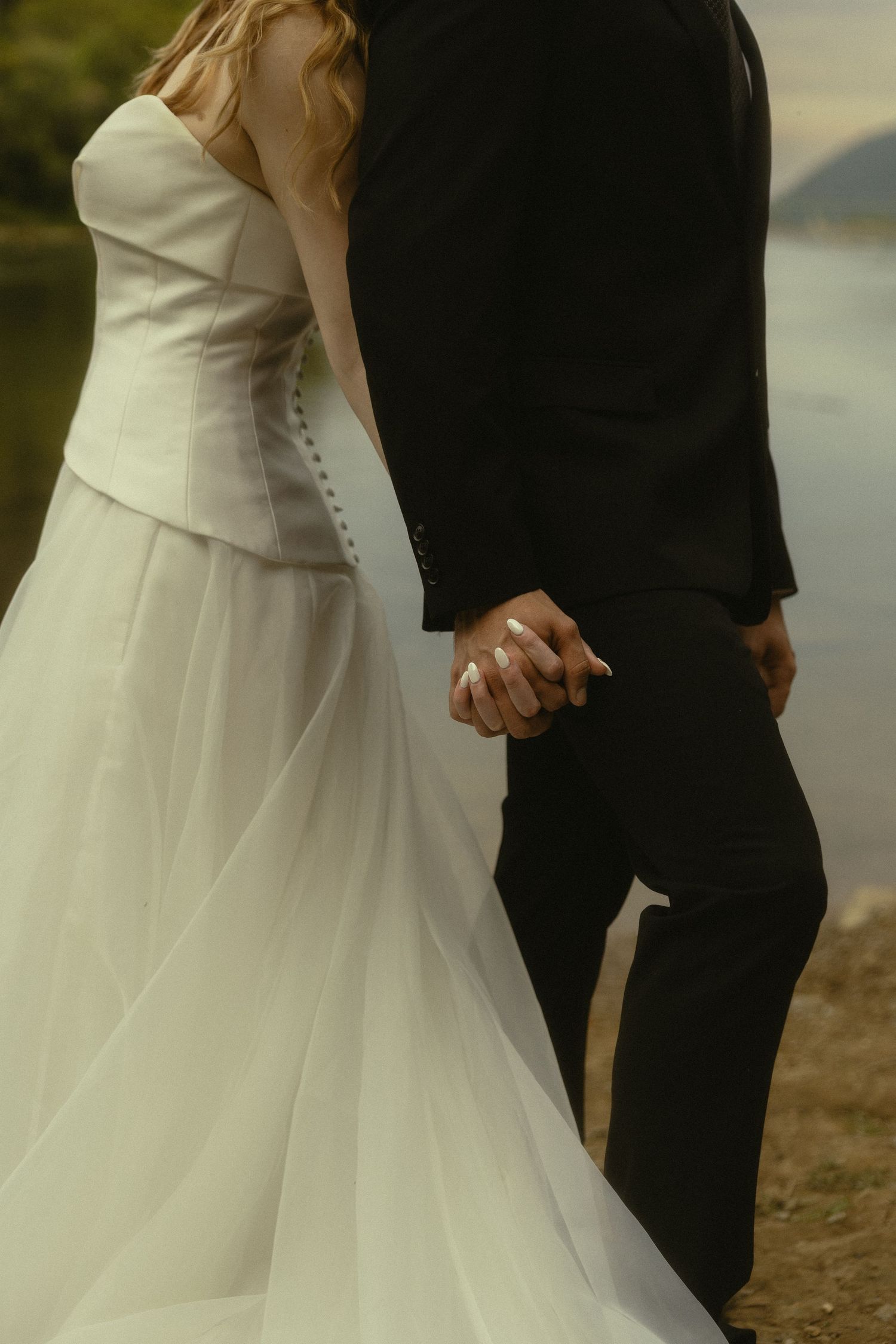 Bride in white gown and groom in black suit holding hands near a lake on their wedding day.