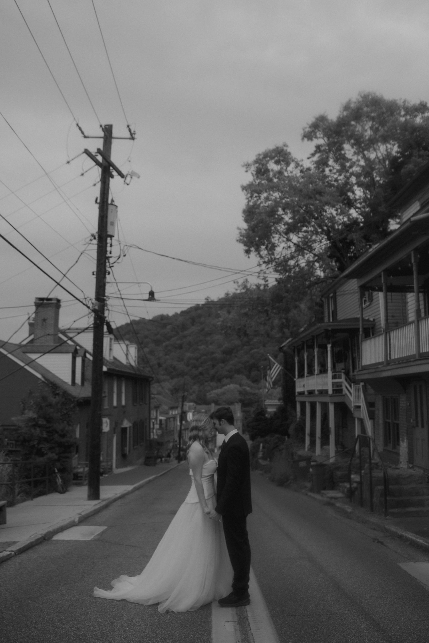 Black and white photo of a bride and groom standing in the middle of a small town street with a forested hill backdrop.