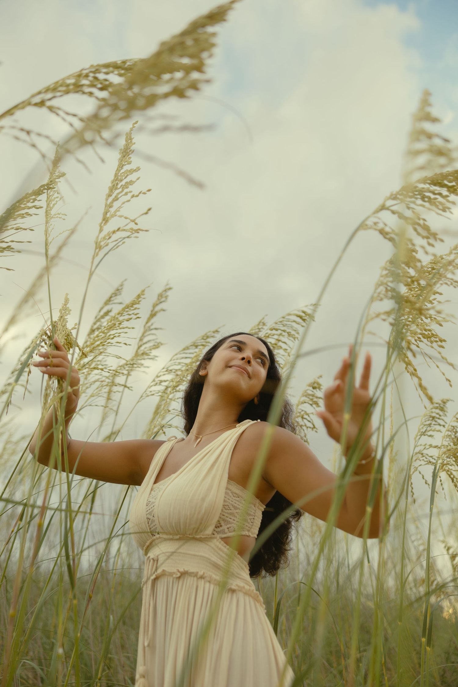 Young woman in cream dress reaching up to touch tall golden grass stalks in a field under a cloudy sky.