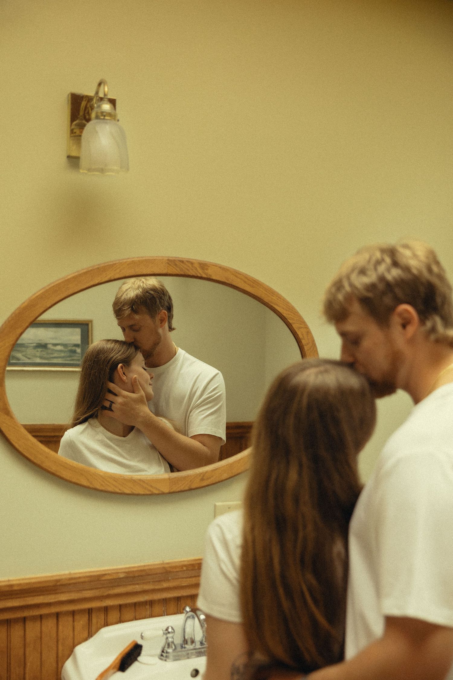 Couple sharing an intimate moment in a bathroom, their embrace reflected in an oval wooden-framed mirror.