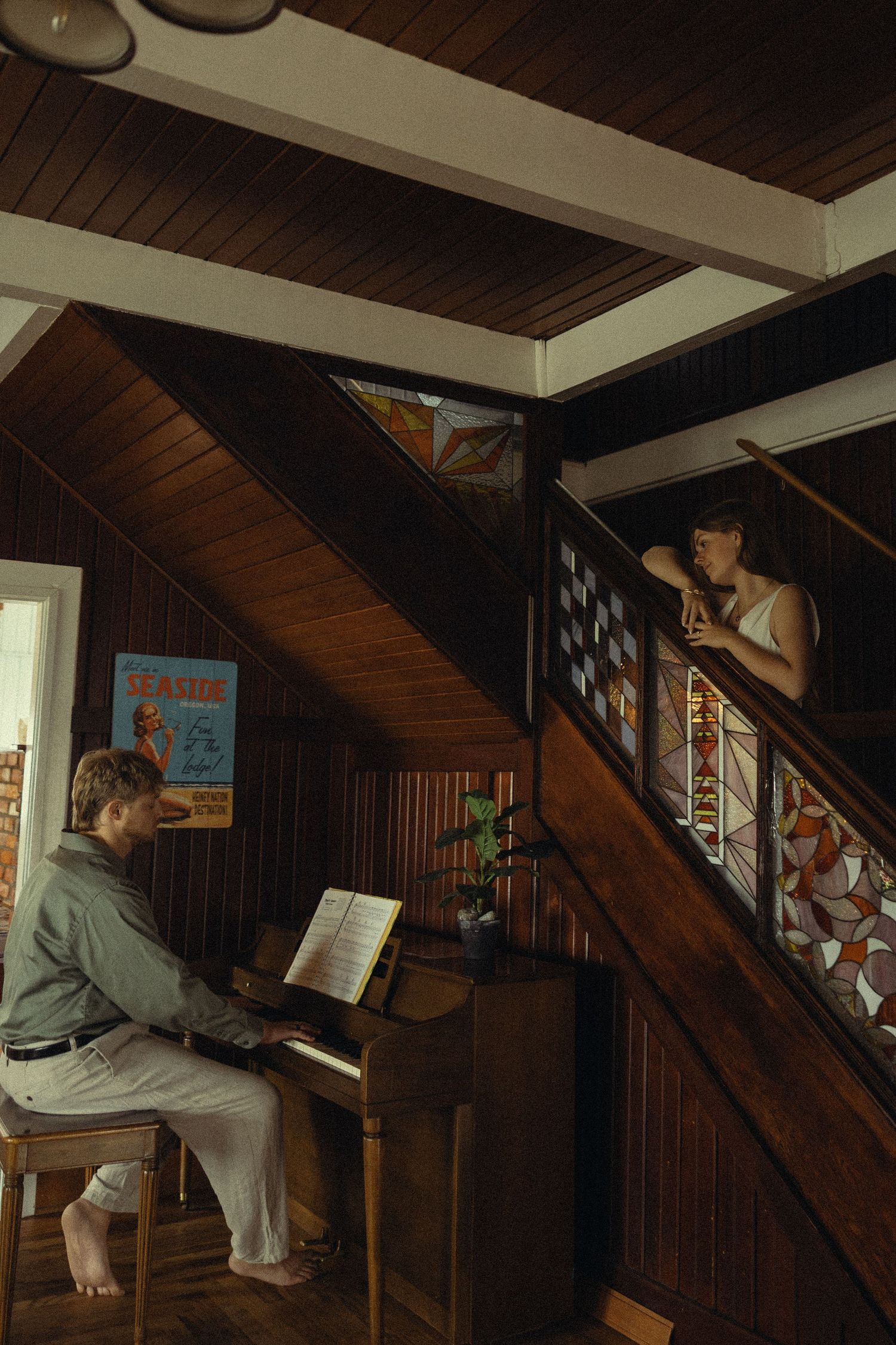 Man playing piano while woman watches from staircase landing in rustic wood-paneled home interior.