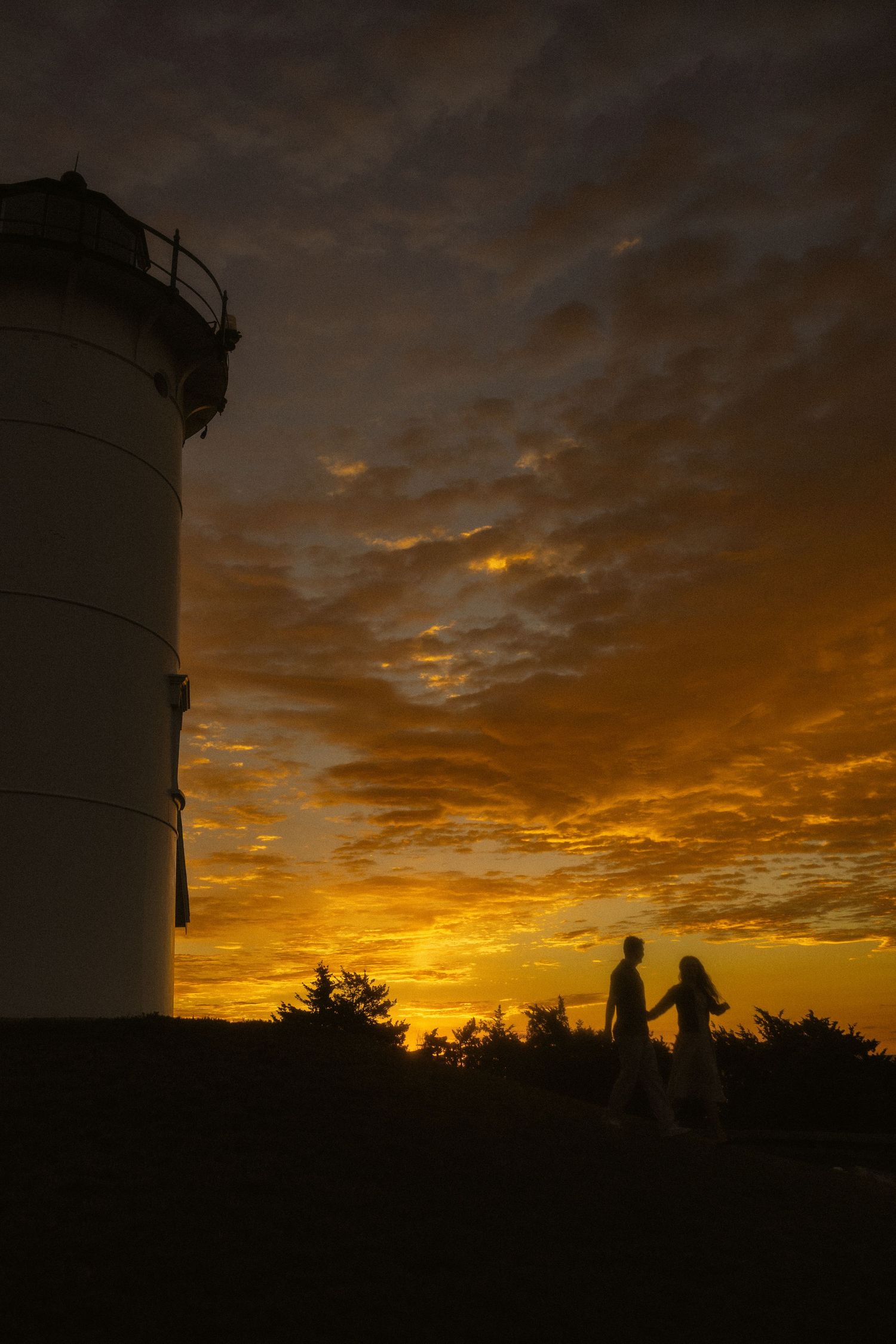 Silhouette of a couple holding hands near a lighthouse against a dramatic golden sunset sky with textured clouds.