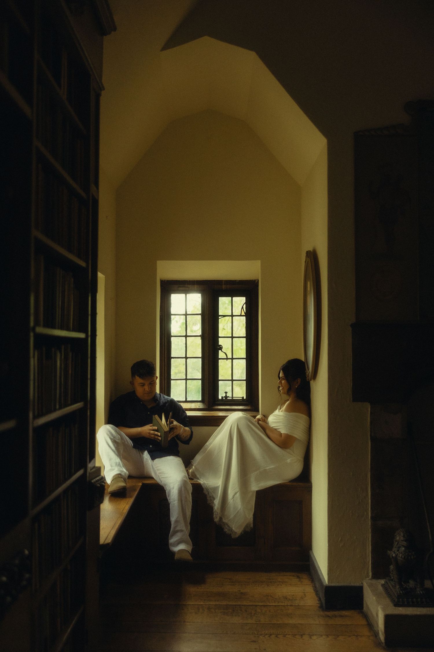 Couple sitting in a cozy library window nook, woman in white dress, man in navy, surrounded by bookshelves.