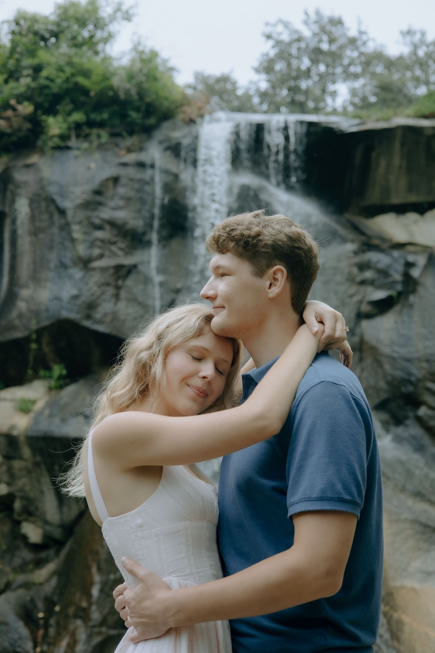 A young couple embraces tenderly in front of a scenic waterfall surrounded by lush greenery.