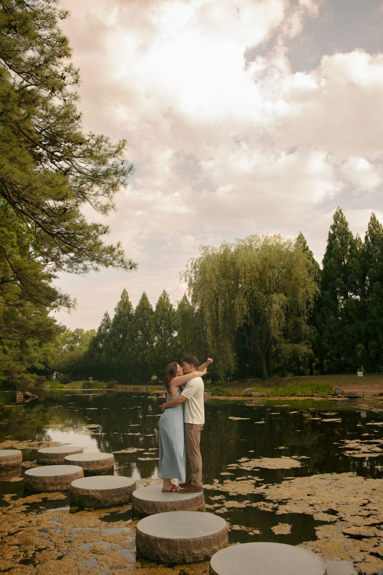 Couple embracing on stepping stones over a serene pond, surrounded by lush trees and soft cloudy sky.