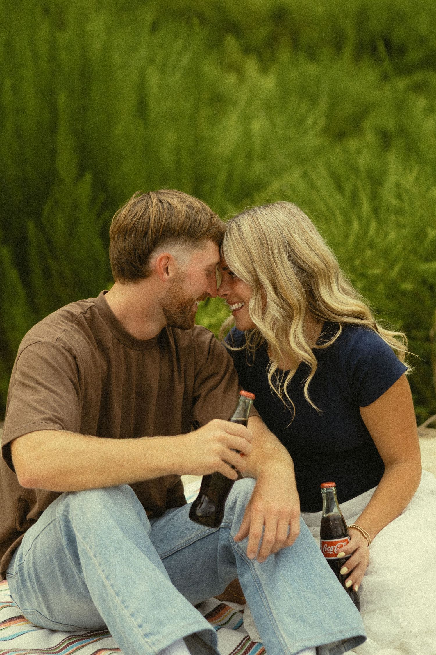 A couple sharing a romantic moment on a blanket outdoors, clinking Coca-Cola bottles while smiling at each other.