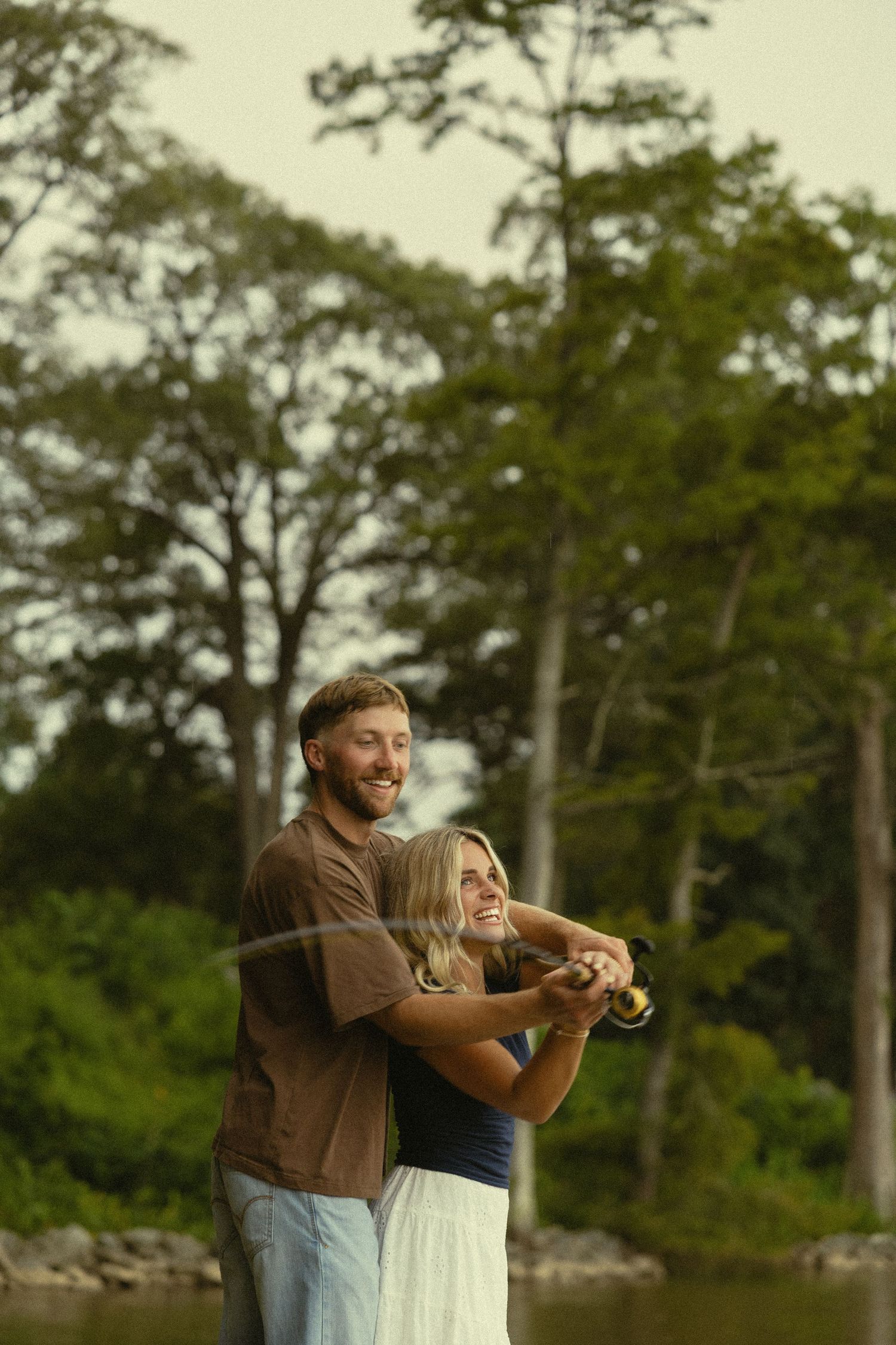 Happy couple fishing together by a lakeside, surrounded by tall green trees in a serene outdoor setting.