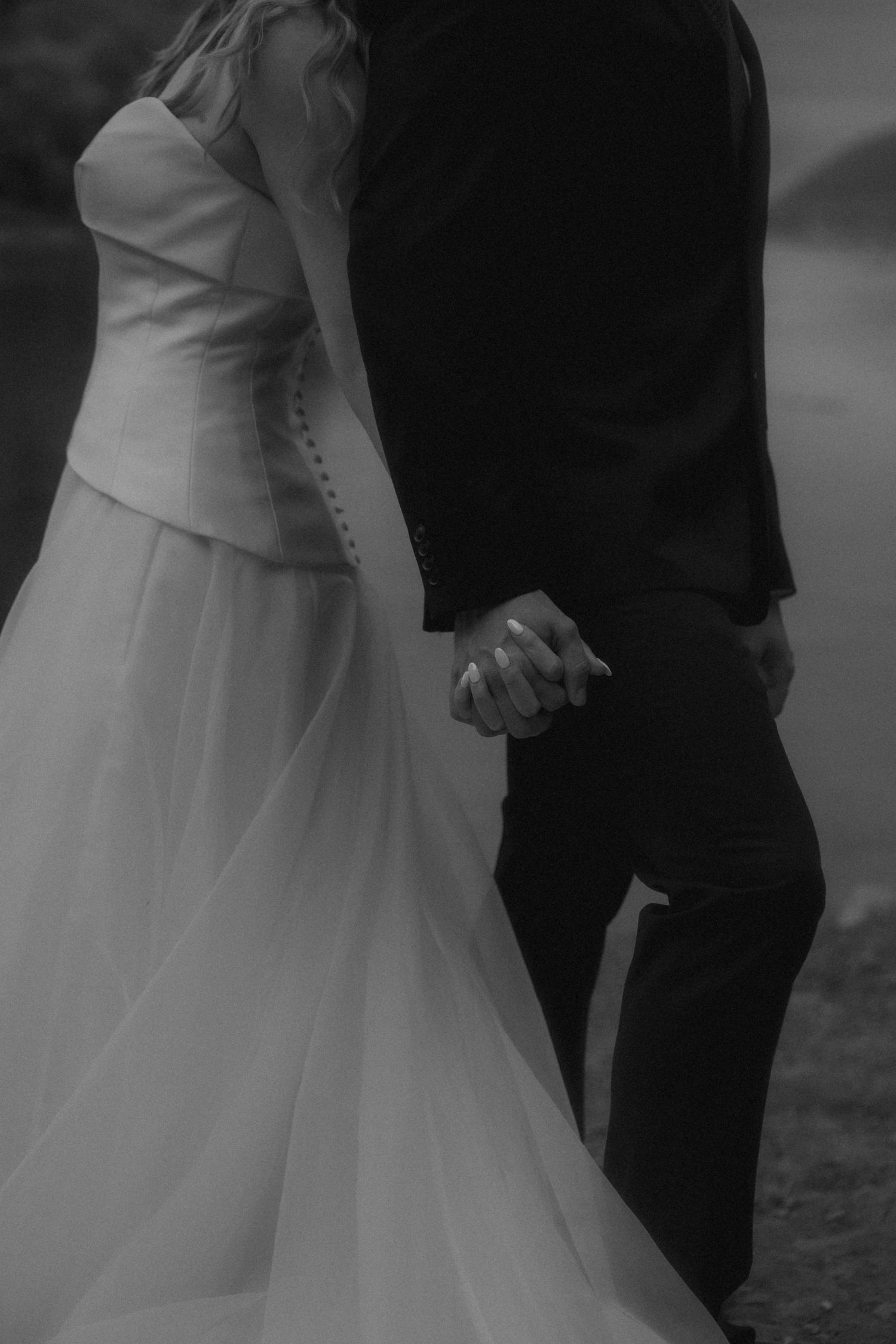 Black and white photo of bride and groom holding hands, bride in elegant white gown and groom in dark suit.