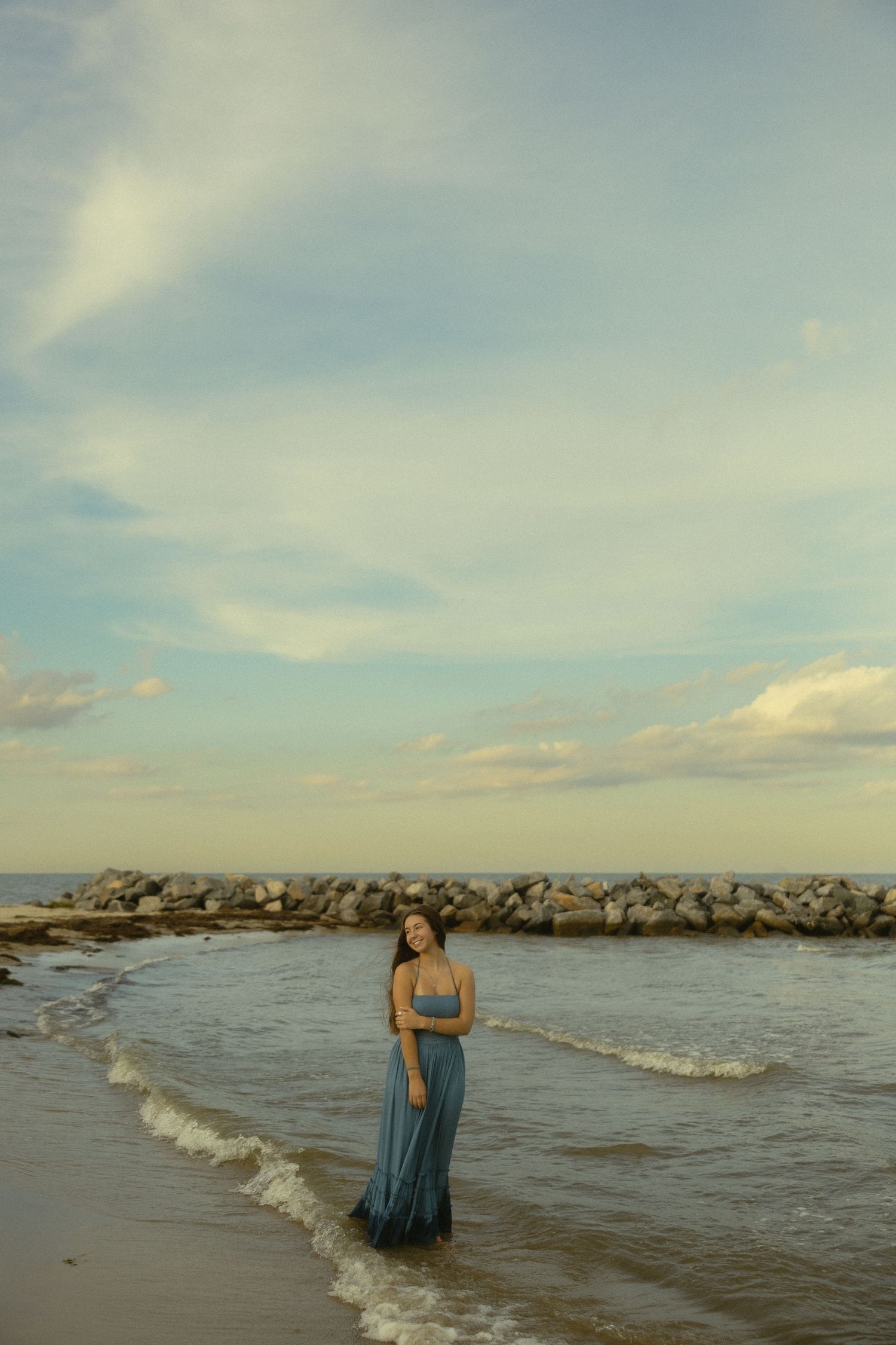 Woman in blue maxi dress standing in ocean waves near rocky shore at golden hour sunset.