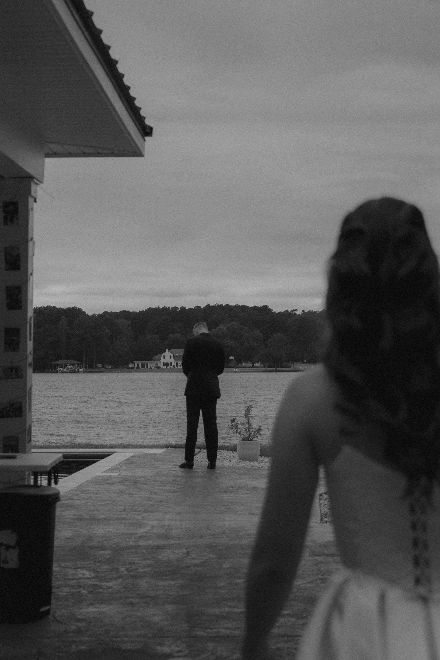 Black and white photo of a bride approaching a groom standing at waterfront edge during an outdoor wedding ceremony.