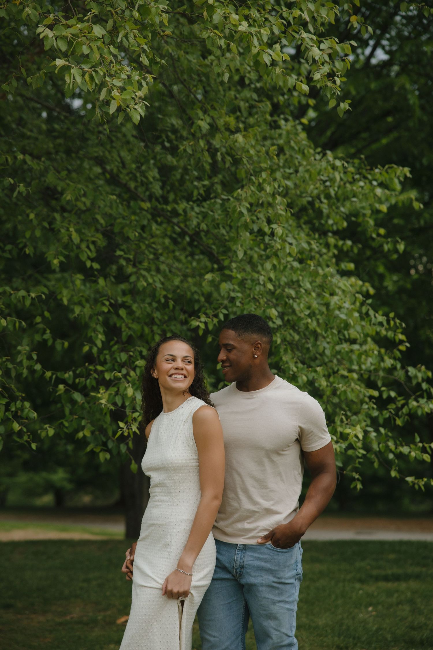 A smiling interracial couple holds hands in a lush green park, woman in white dress, man in beige shirt and jeans.