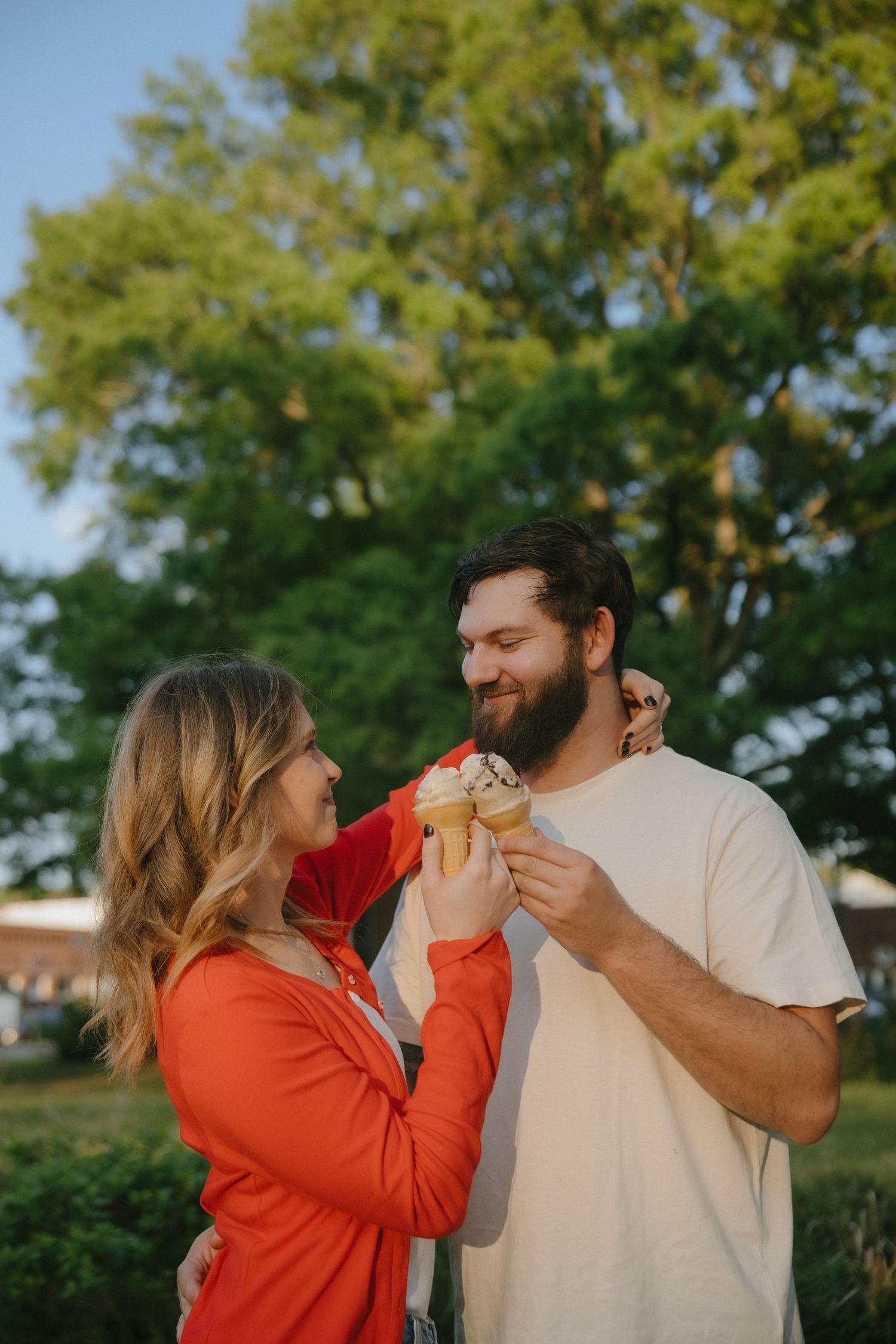A couple sharing ice cream cones outdoors, smiling together near lush green trees on a sunny day.
