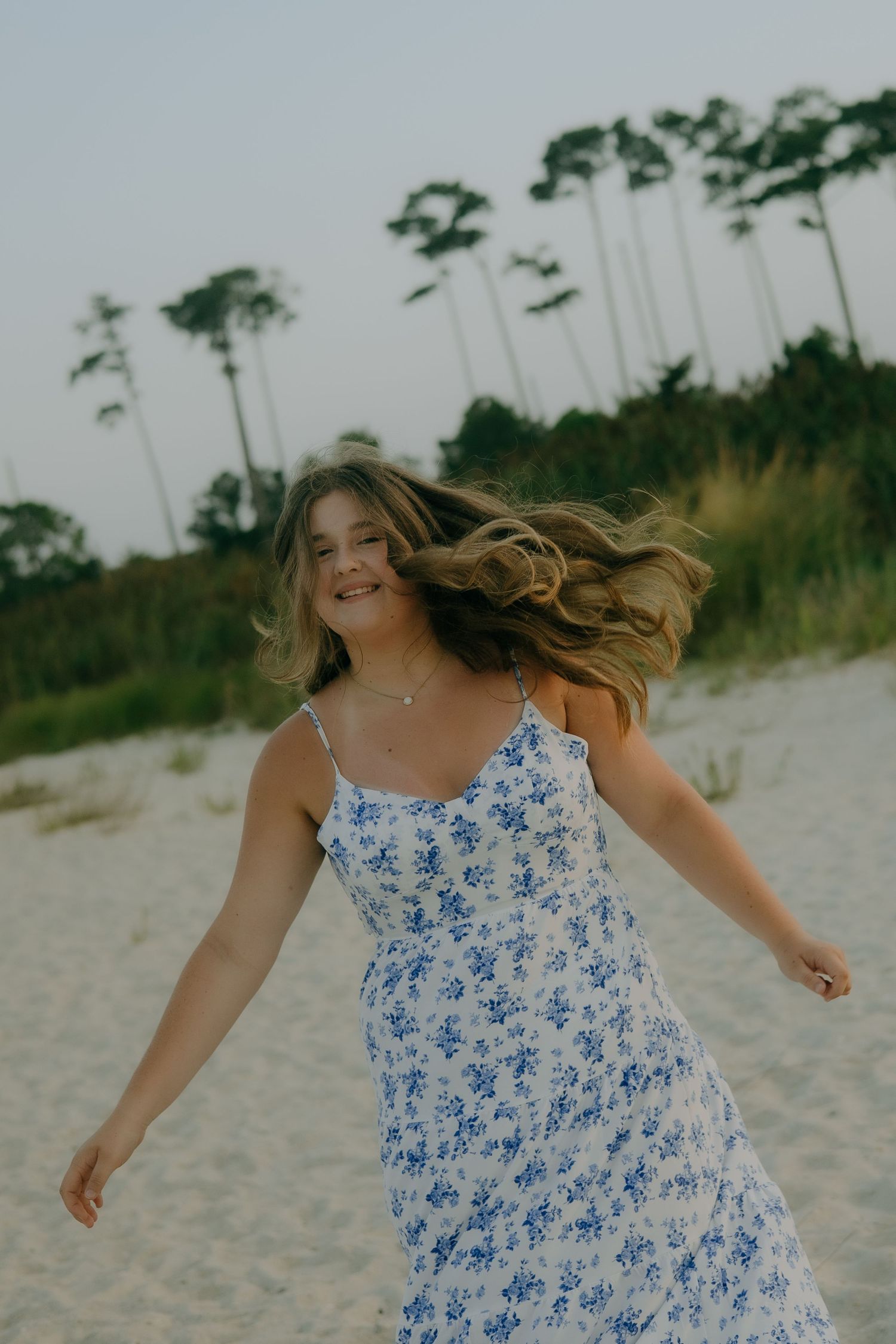 A young woman in a blue floral sundress twirls on a sandy beach with tall pine trees in the background.