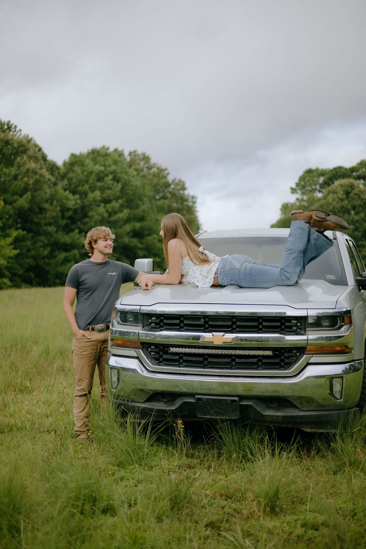 Two people pose playfully near a modern Chevrolet Silverado truck in a green field.