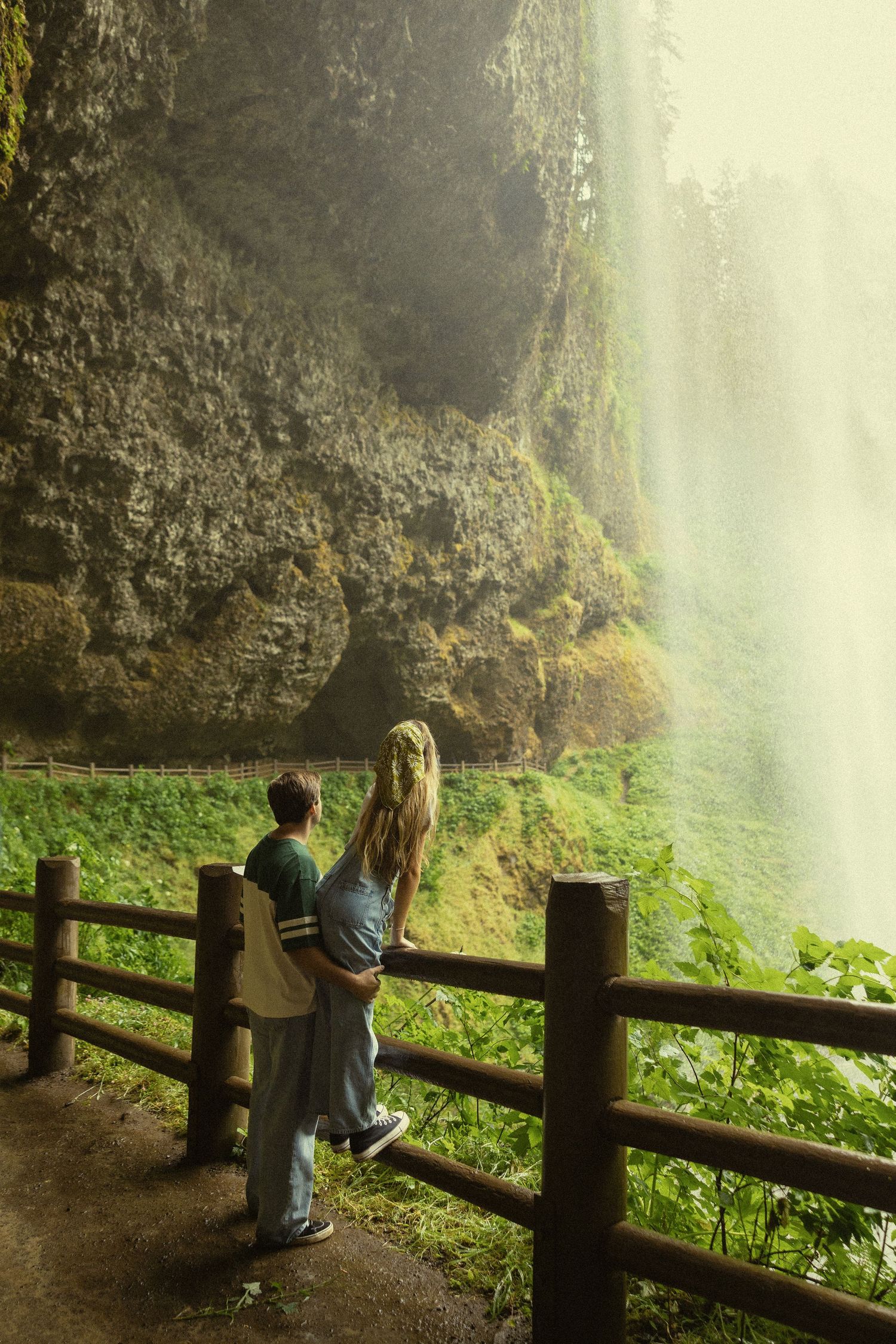 Couple standing at wooden fence viewpoint admiring a majestic waterfall cascading down mossy cliffs in a lush green forest.