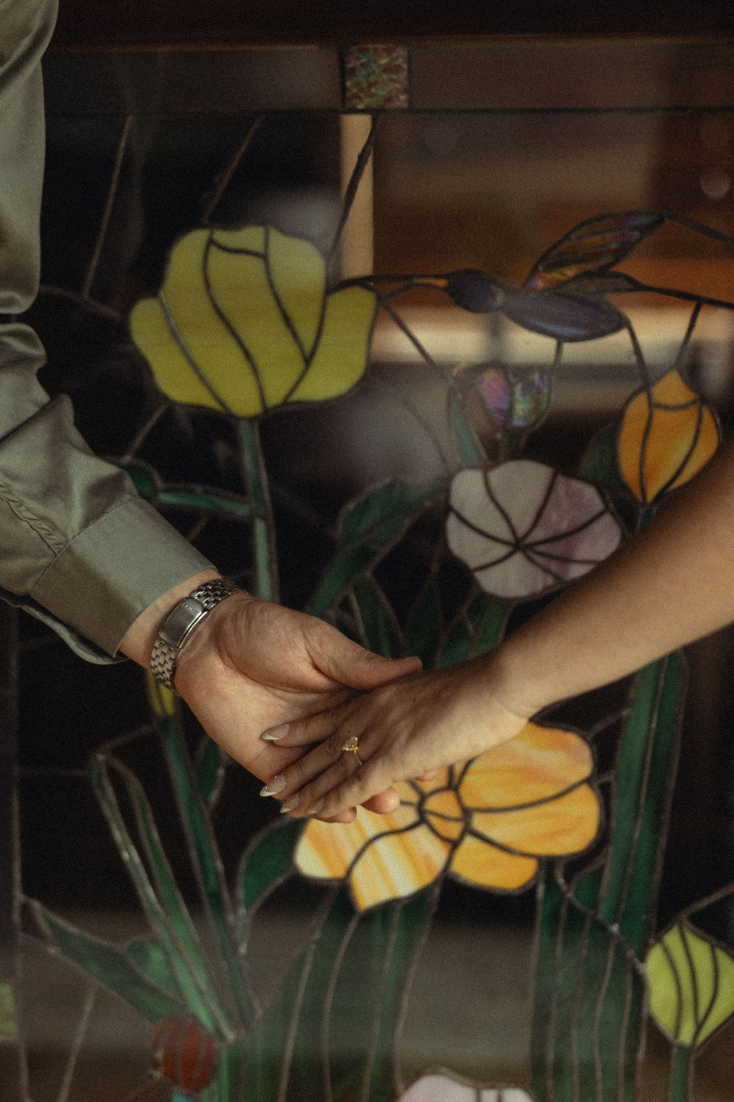 Two people holding hands in front of a colorful stained glass window with floral designs.