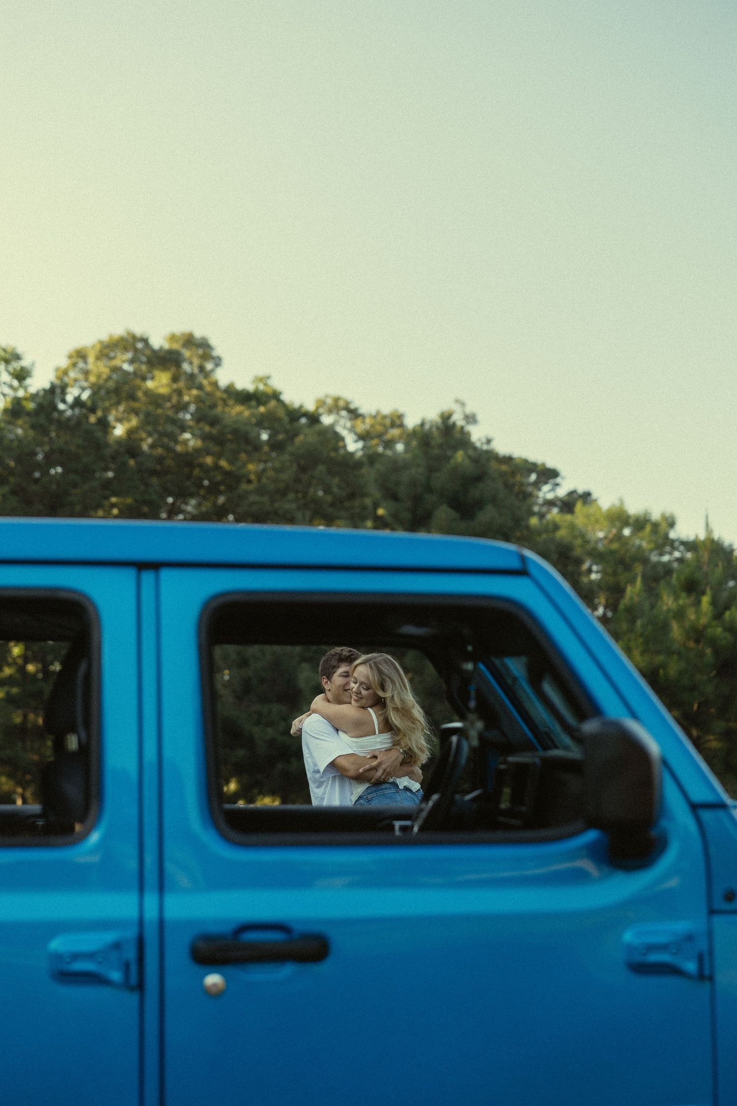 Couple embracing romantically reflected in the window of a blue SUV with green trees in the background.