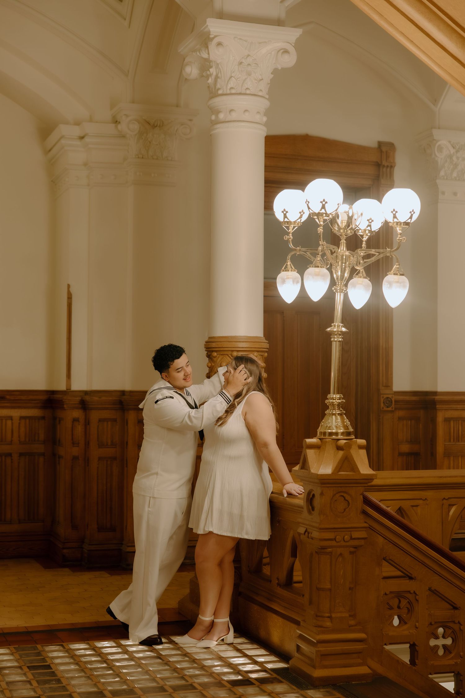 Couple in white outfits share a tender moment in an elegant hall with ornate columns and a gold candelabra.