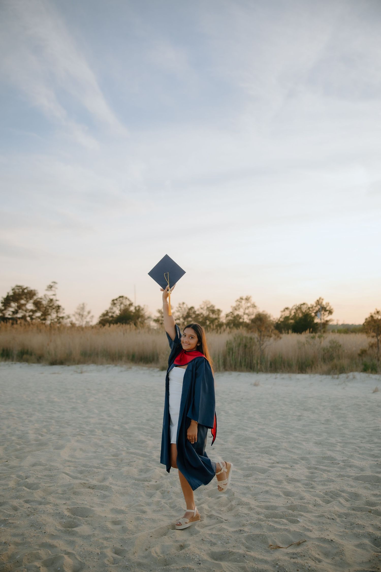 Young woman in graduation gown and cap celebrating on sandy beach at sunset.