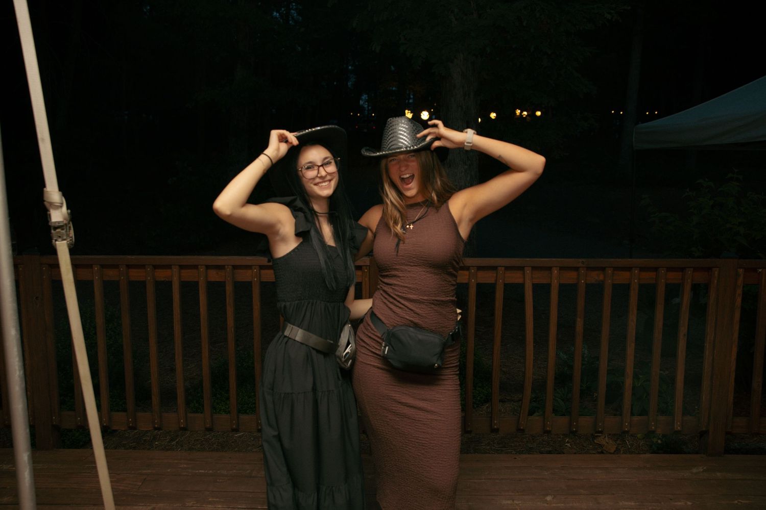 Two women in cowboy hats posing on a wooden deck at night, smiling and touching their hat brims.