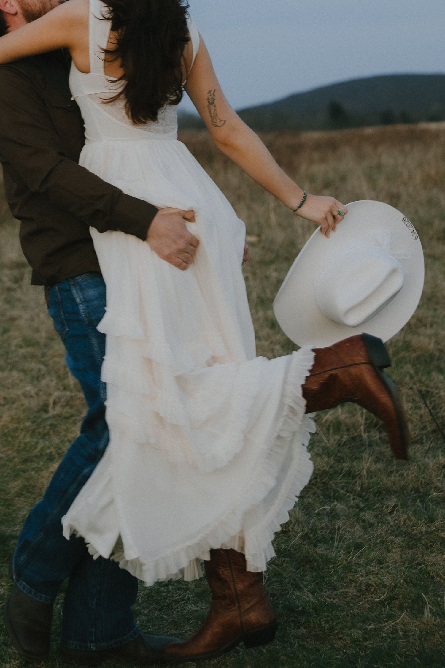 Bride in ruffled white dress and cowboy boots dances with groom in a rustic field.