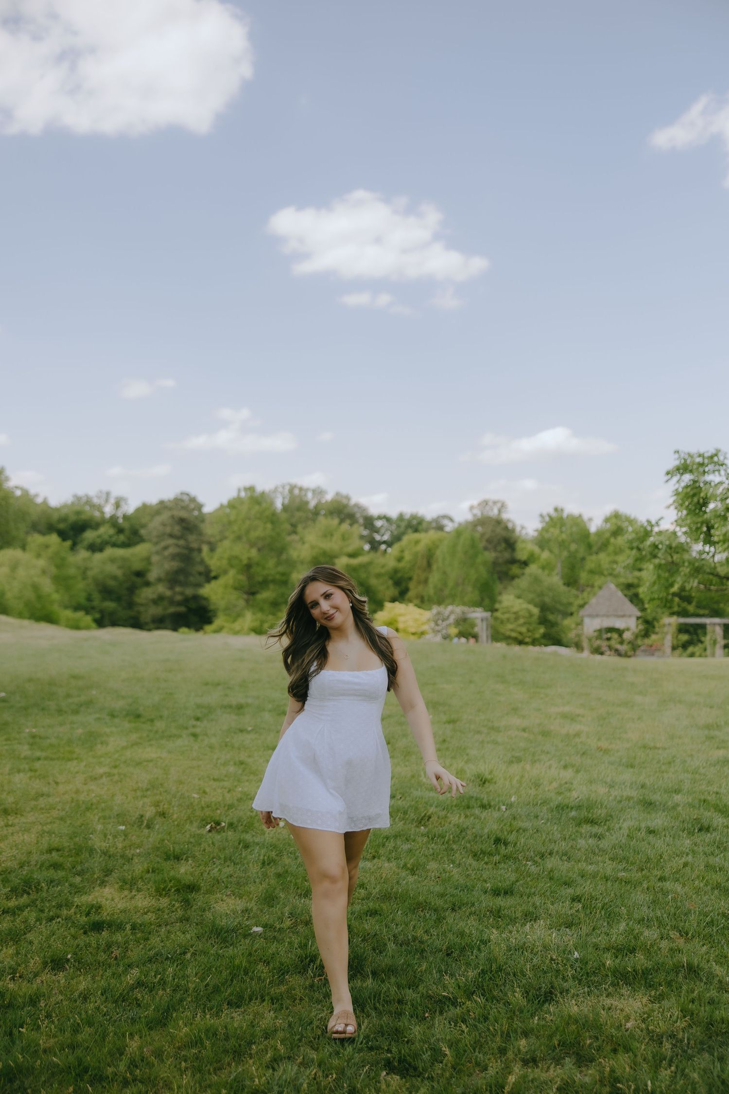 Young woman in white sundress walking on lush green lawn with trees and blue sky in background.