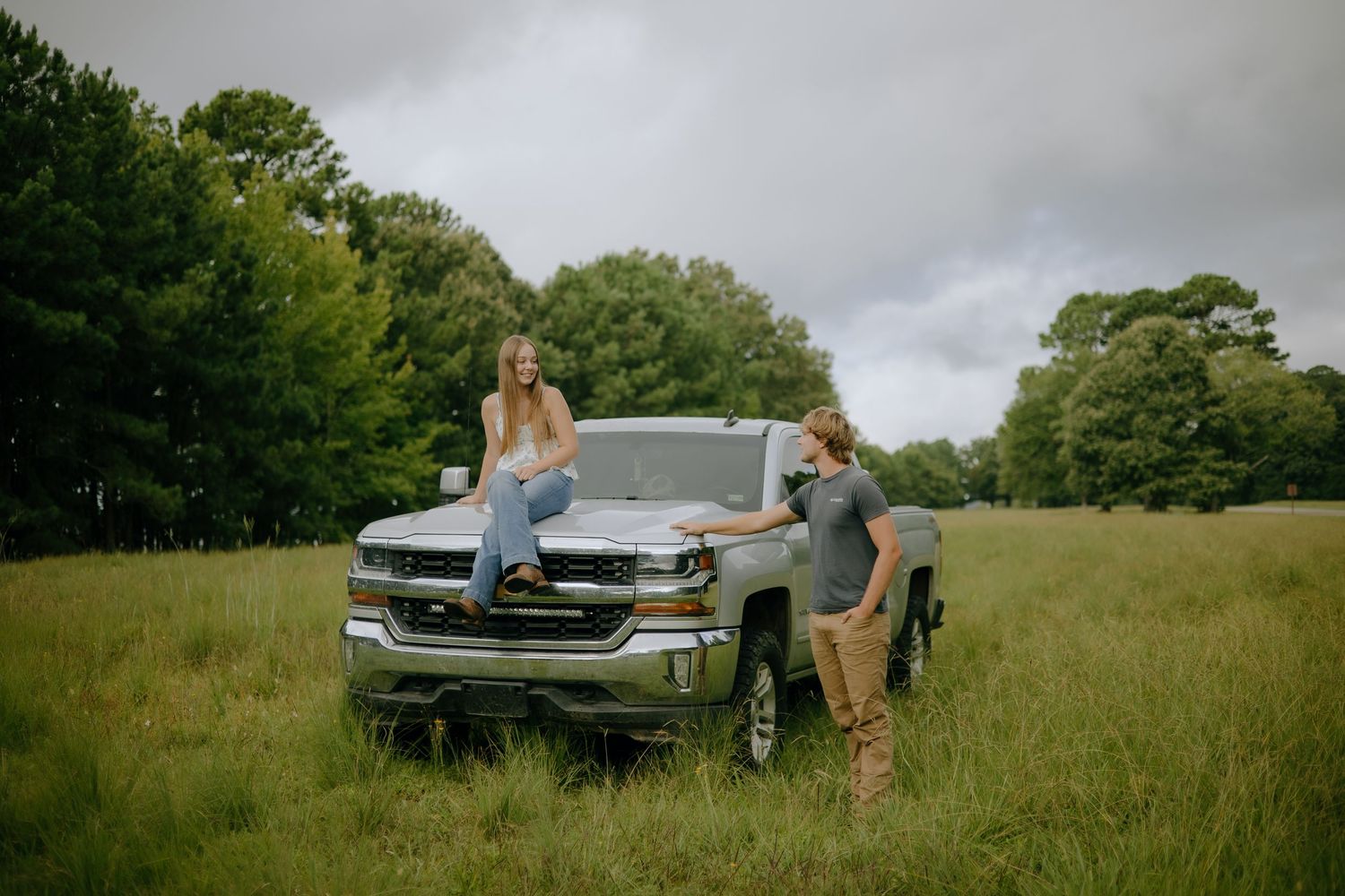 People gather around a silver pickup truck parked in a grassy field on an overcast day.