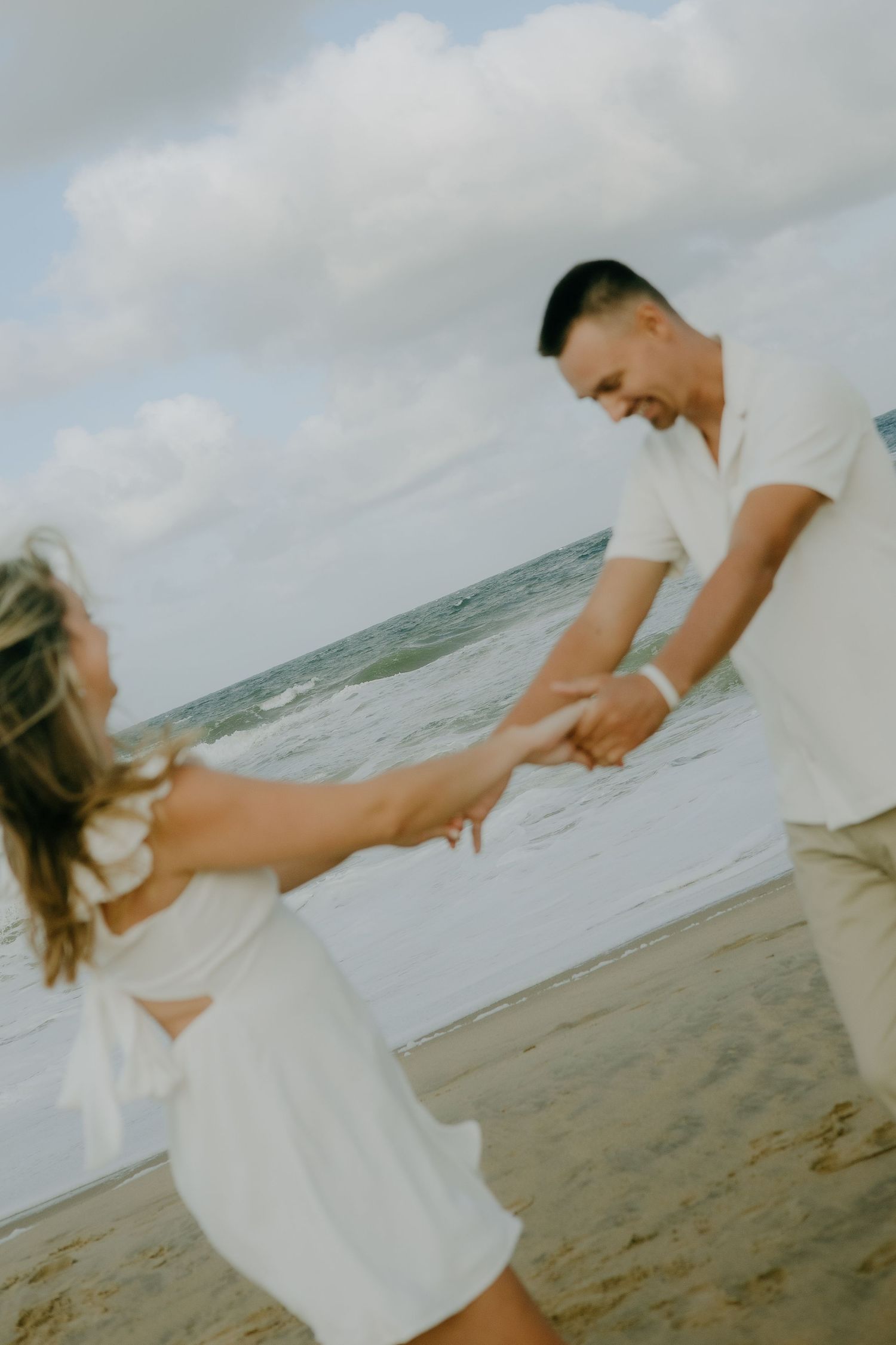 A couple dances and spins together on a sandy beach with ocean waves behind them.
