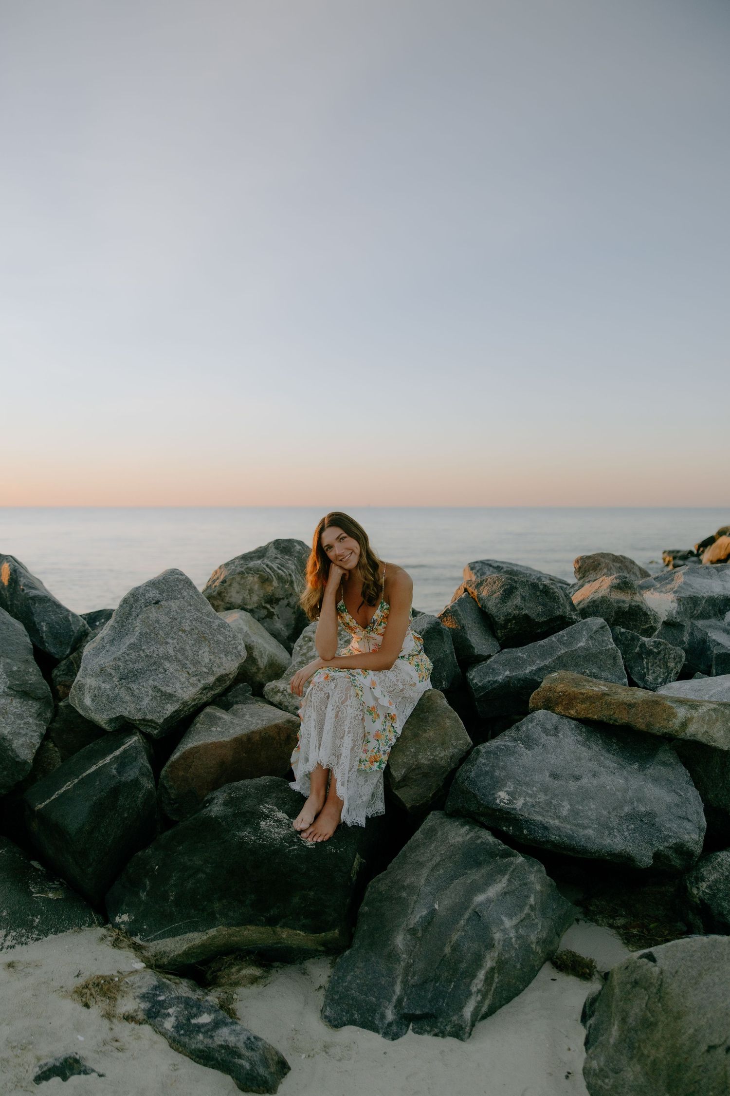Woman in floral dress sitting on rocks at beach during sunset with calm ocean in background.