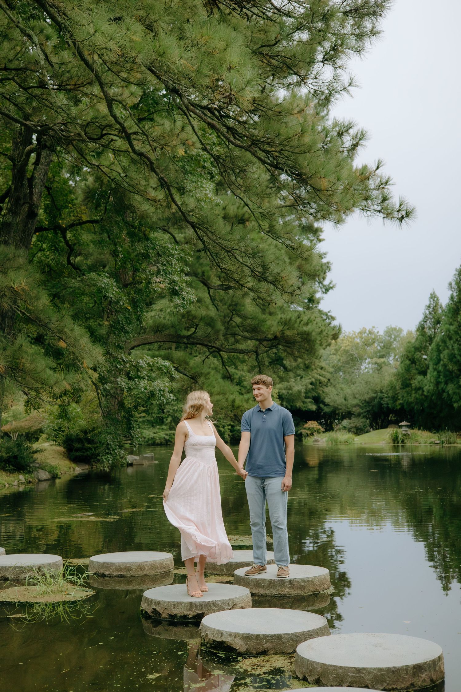 A couple walks along stepping stones across a tranquil pond surrounded by overhanging pine trees on a peaceful afternoon.