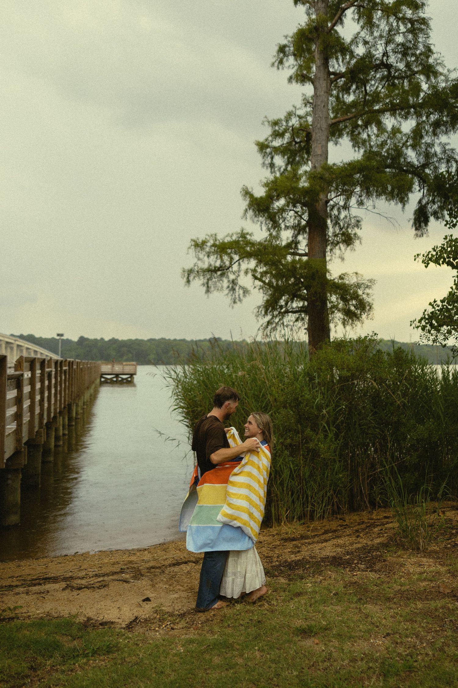 Couple embracing by a lakeside dock surrounded by tall reeds and cypress trees on a cloudy day.