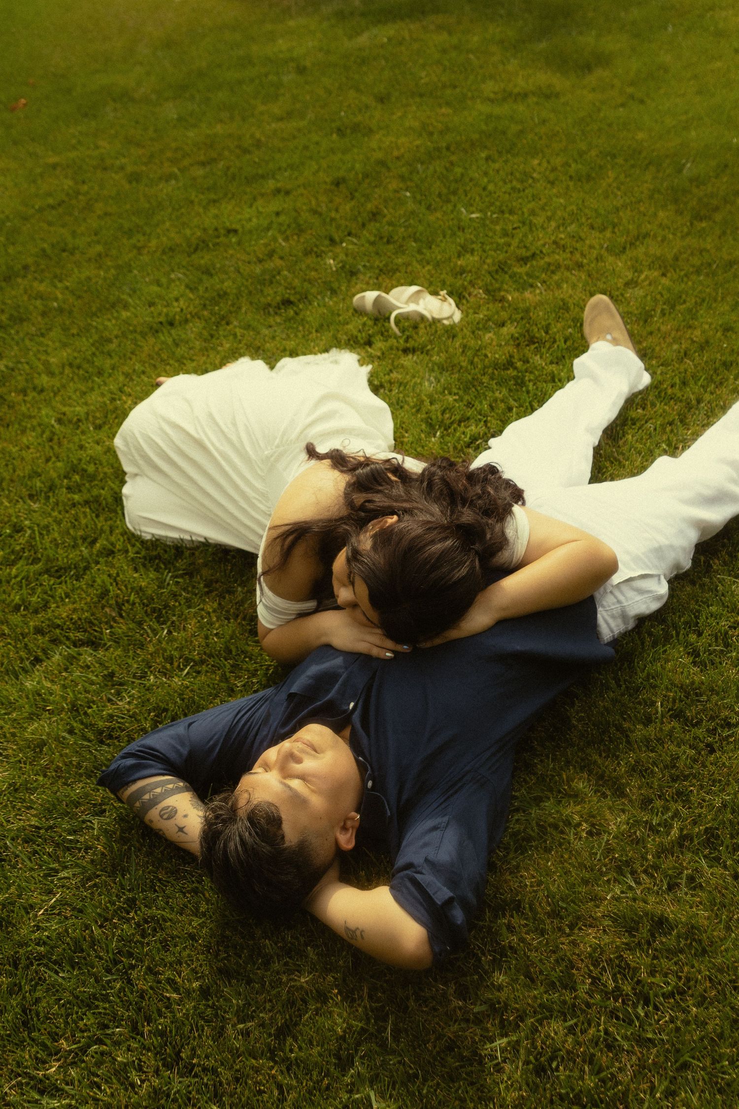 Couple lying on green grass, woman in white dress resting head on man in navy shirt, sharing a tender romantic moment.