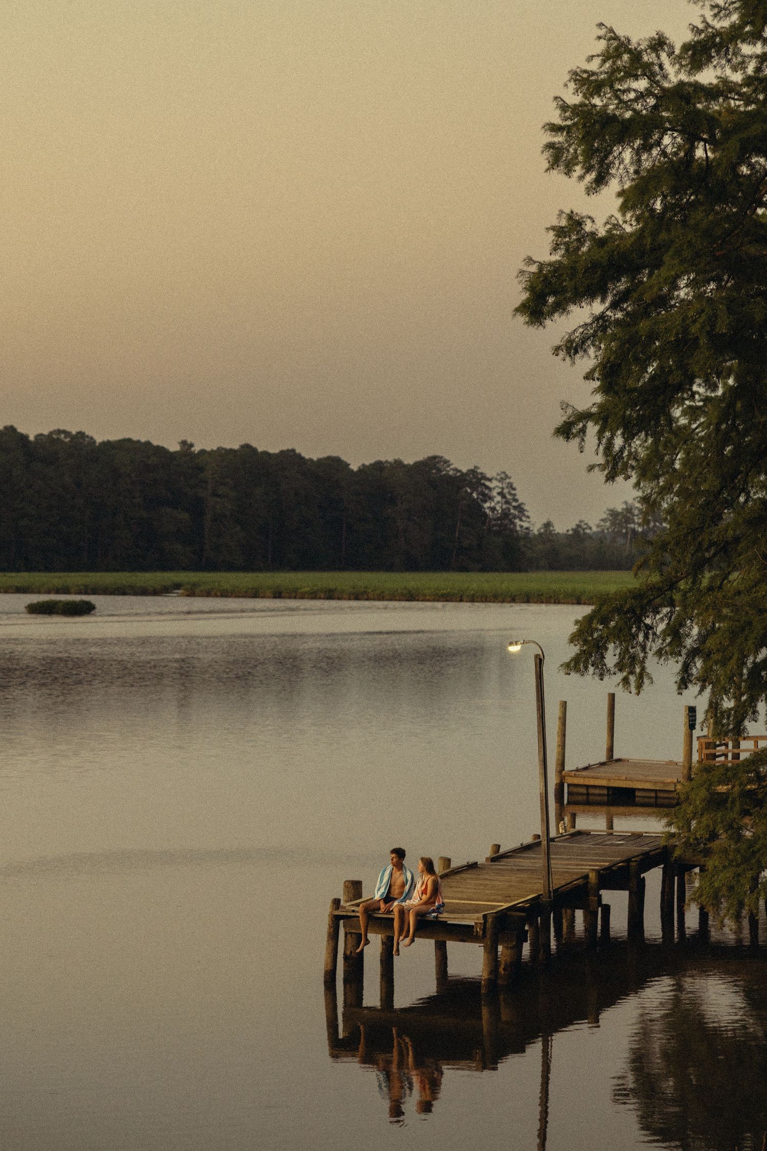 Two people sit on a wooden dock at dusk, reflecting on a calm lake surrounded by lush trees.