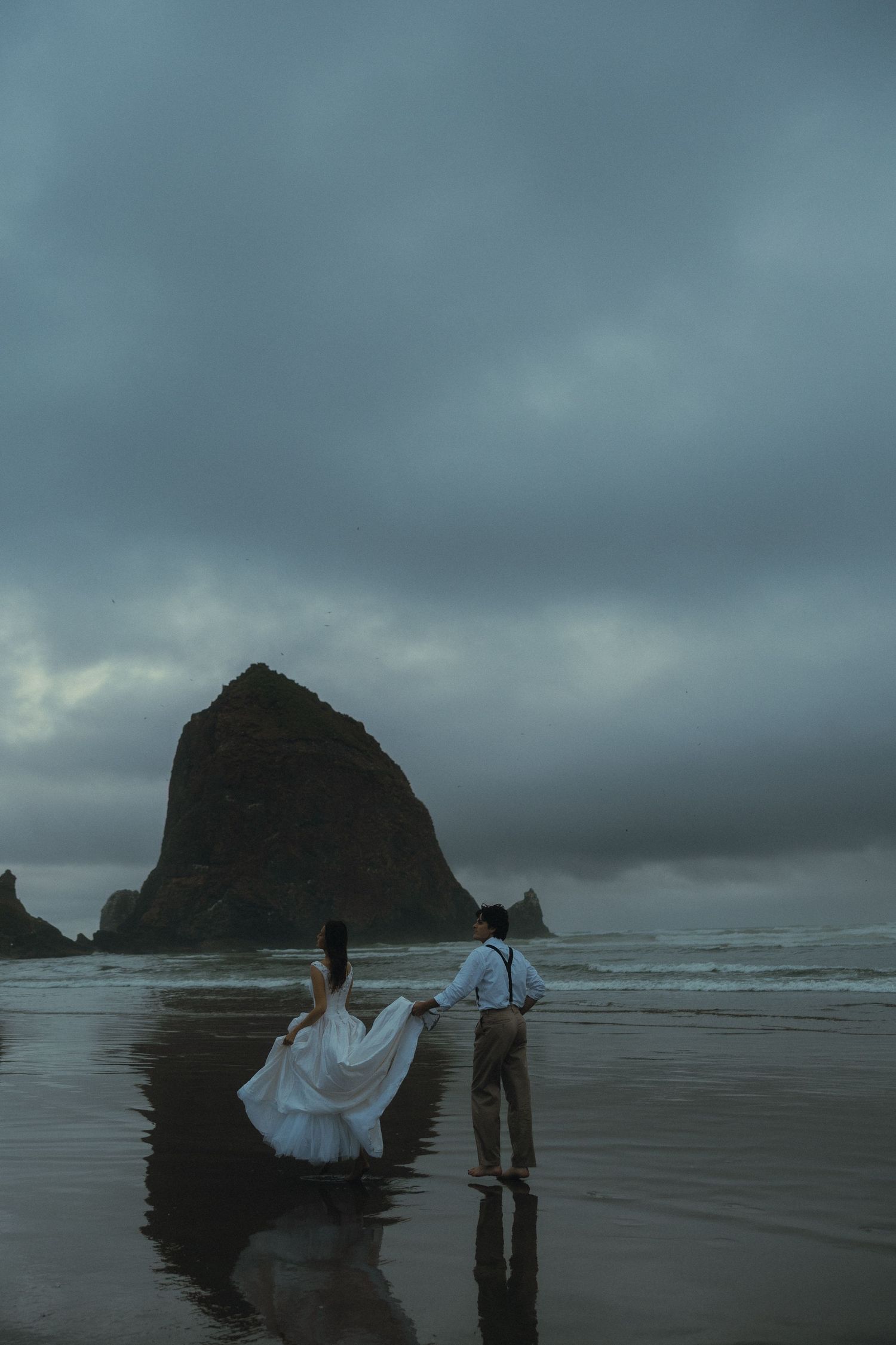 Wedding couple holding hands on a dramatic beach with iconic Haystack Rock in the background at dusk.