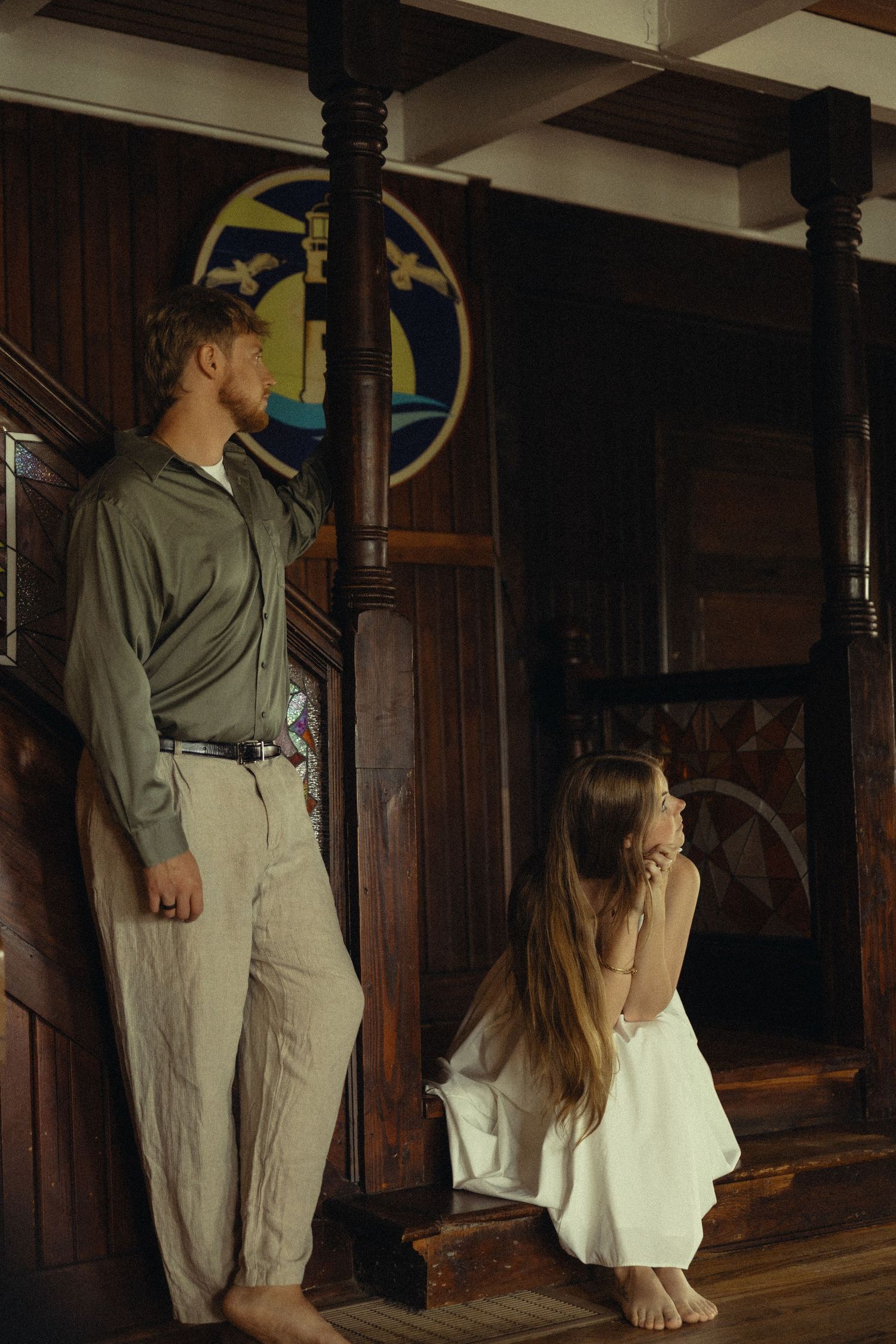 Man in green shirt stands beside a wooden pillar as a woman in white dress sits on steps in a rustic interior.