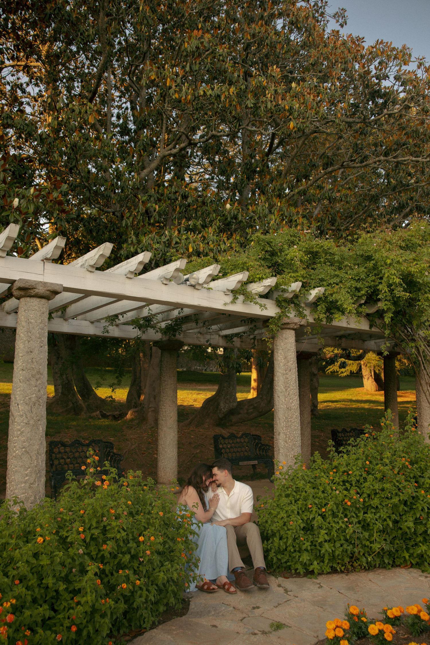 Couple sharing a romantic moment under a white pergola surrounded by lush greenery and orange flowers in a garden.