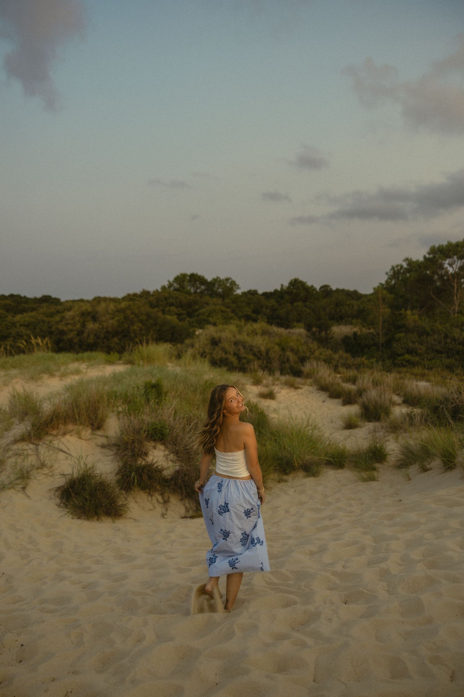 Woman in white top and blue floral skirt walking on sandy dunes with green trees under a twilight sky.