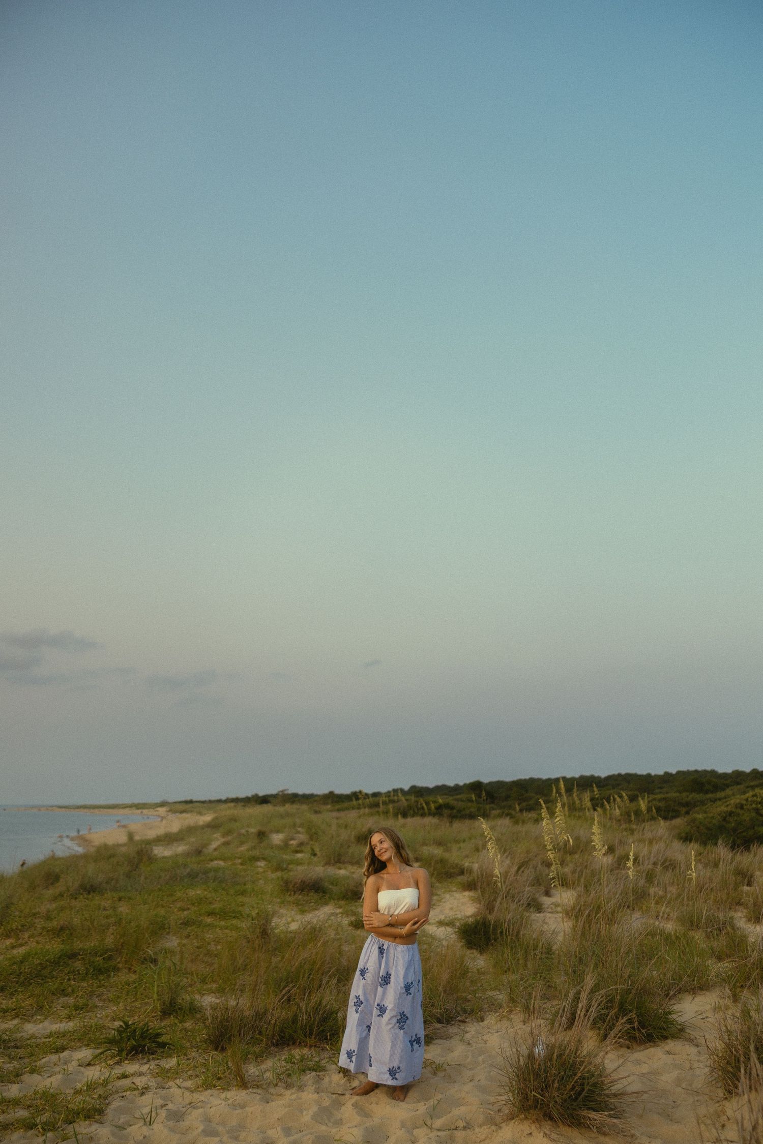 Woman in white top and blue floral skirt standing on sandy beach dunes at dusk with calm ocean in background.