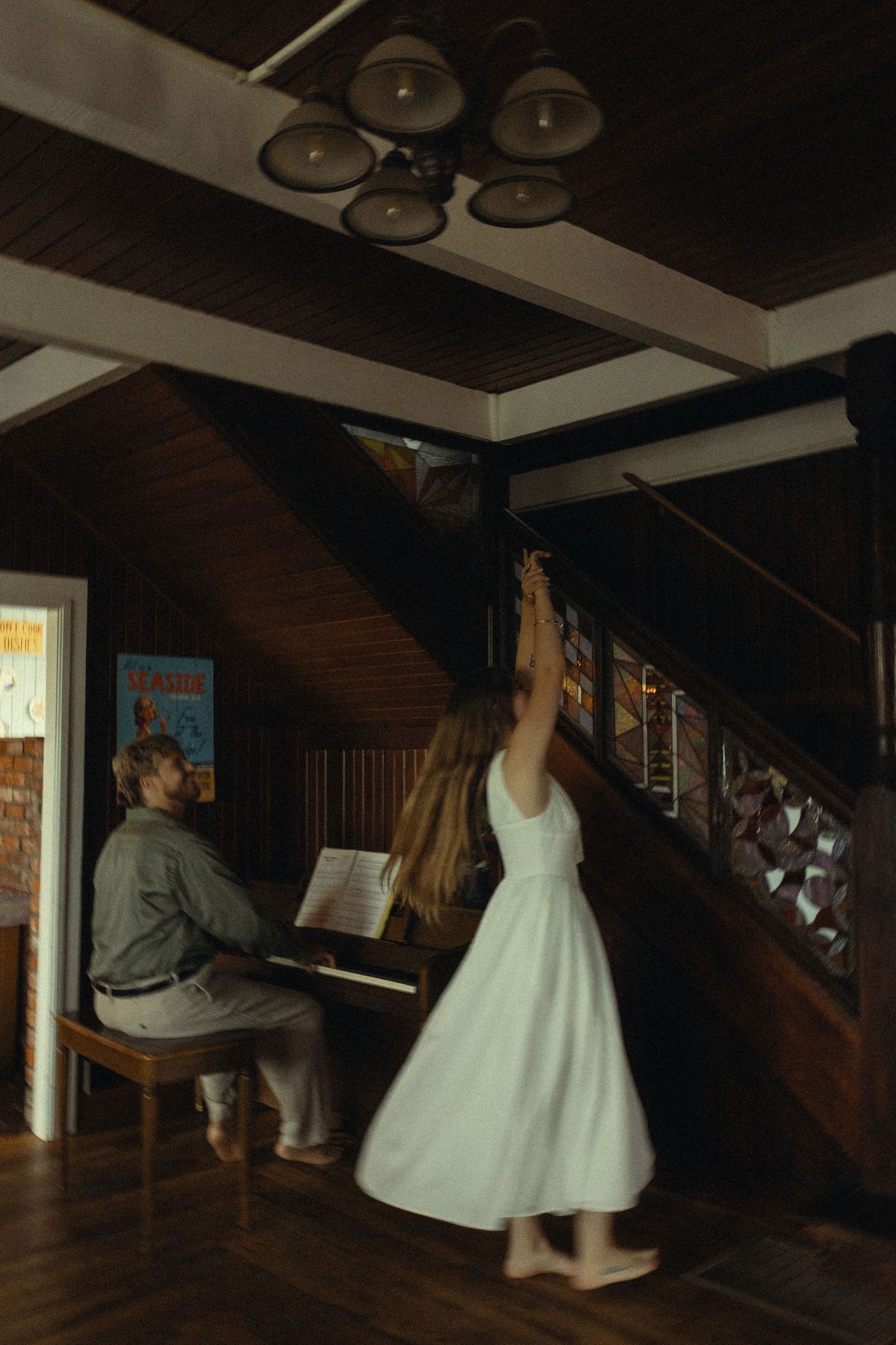 Woman in white dress dancing near stairs while man plays piano in rustic wood-paneled room.