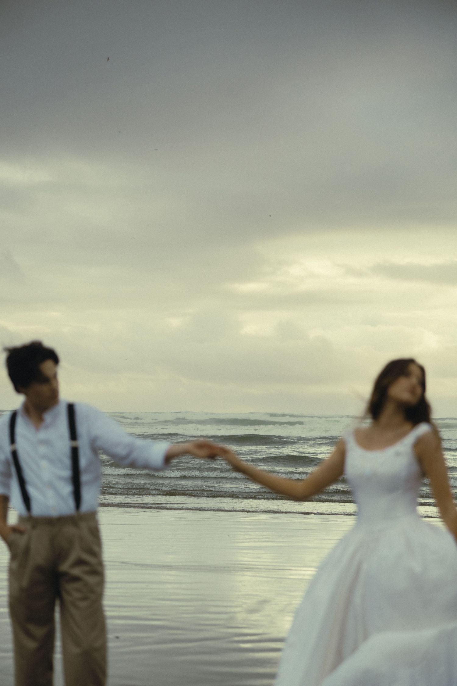Couple in wedding attire holding hands on a misty beach, bride in white gown and groom in suspenders.