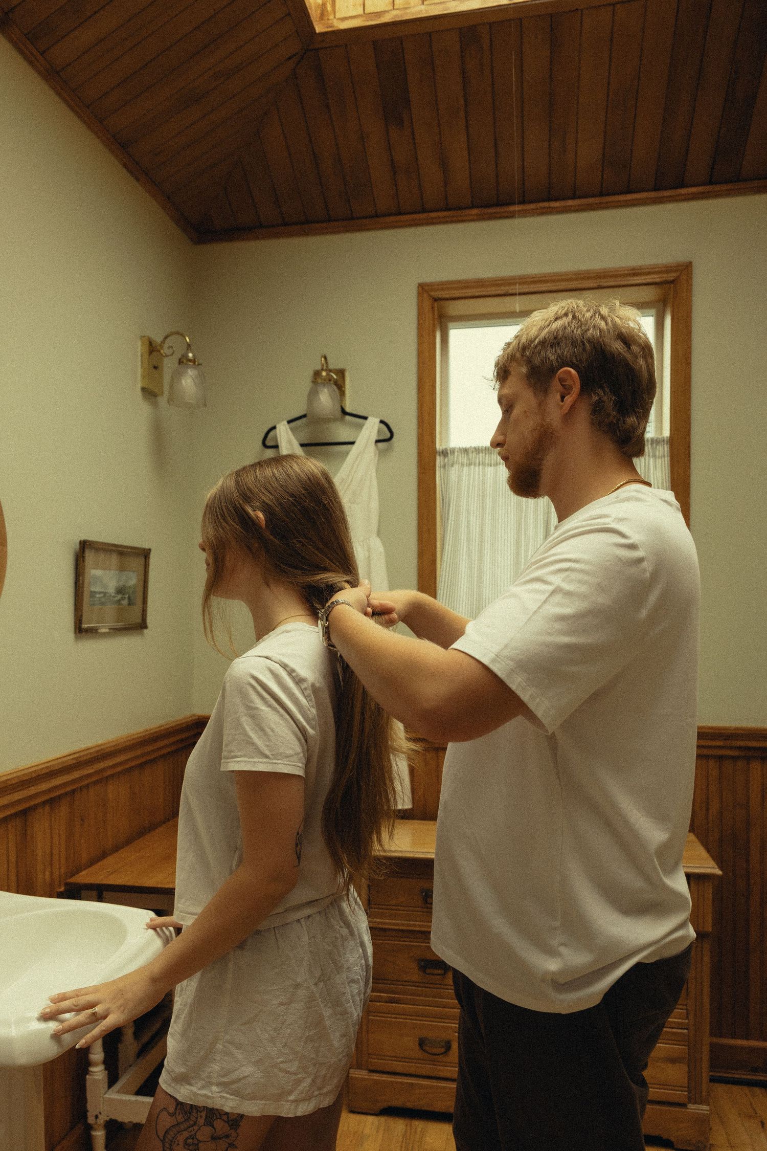 A man braids a woman's long hair in a rustic wooden bathroom with a white dress on a mannequin in the background.